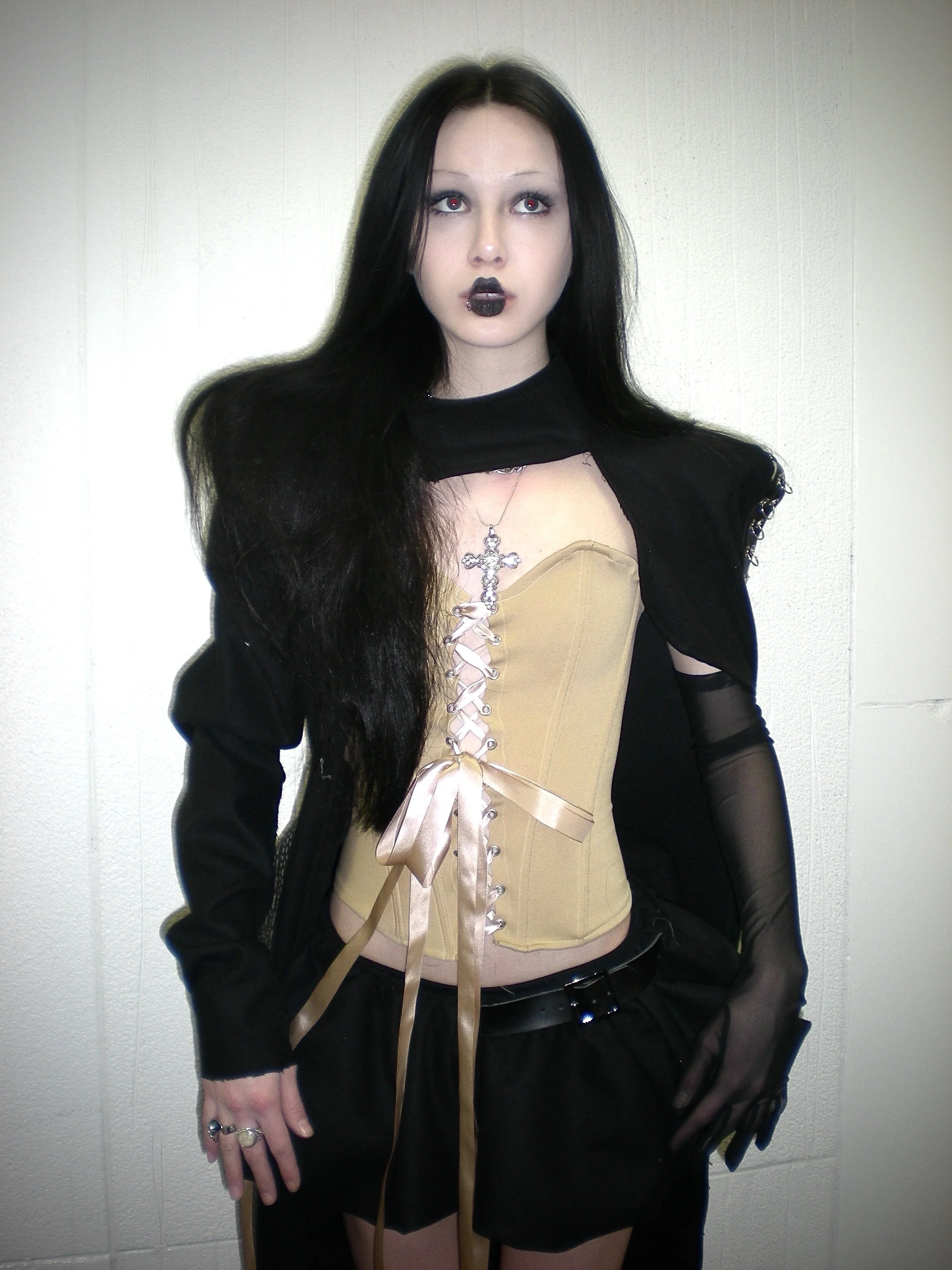 A young woman with long black hair, pale skin, and dark makeup wearing gothic attire, including a beige corset with ribbon lacing, black sheer gloves, black shorts, and a black jacket, standing against a plain light-colored wall.