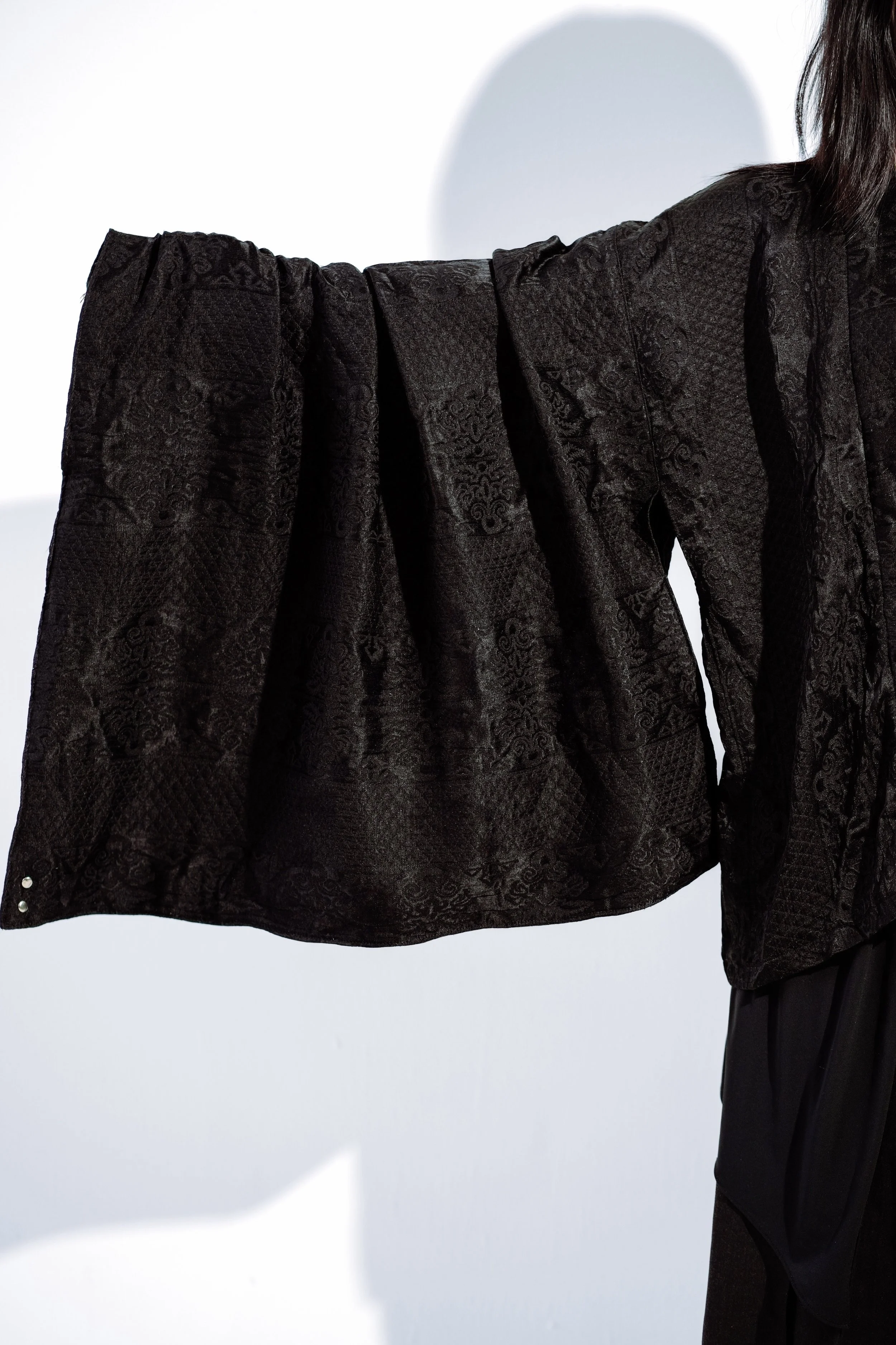 Close-up of a person wearing a black textured, patterned garment with voluminous sleeves against a white background.