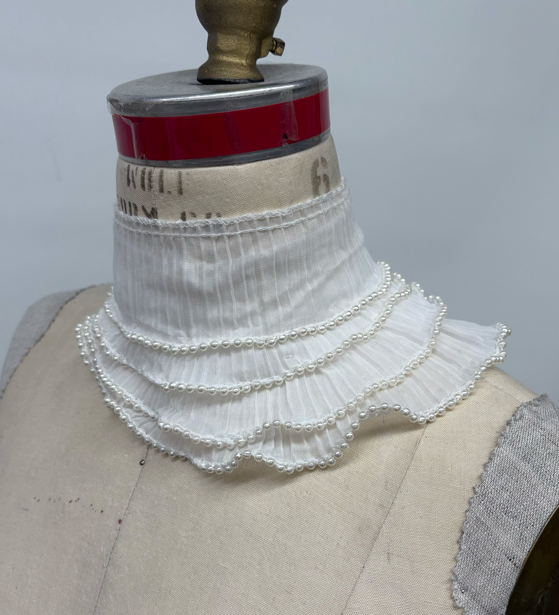 A white, layered, pearl-embellished fabric collar displayed on a mannequin bust.