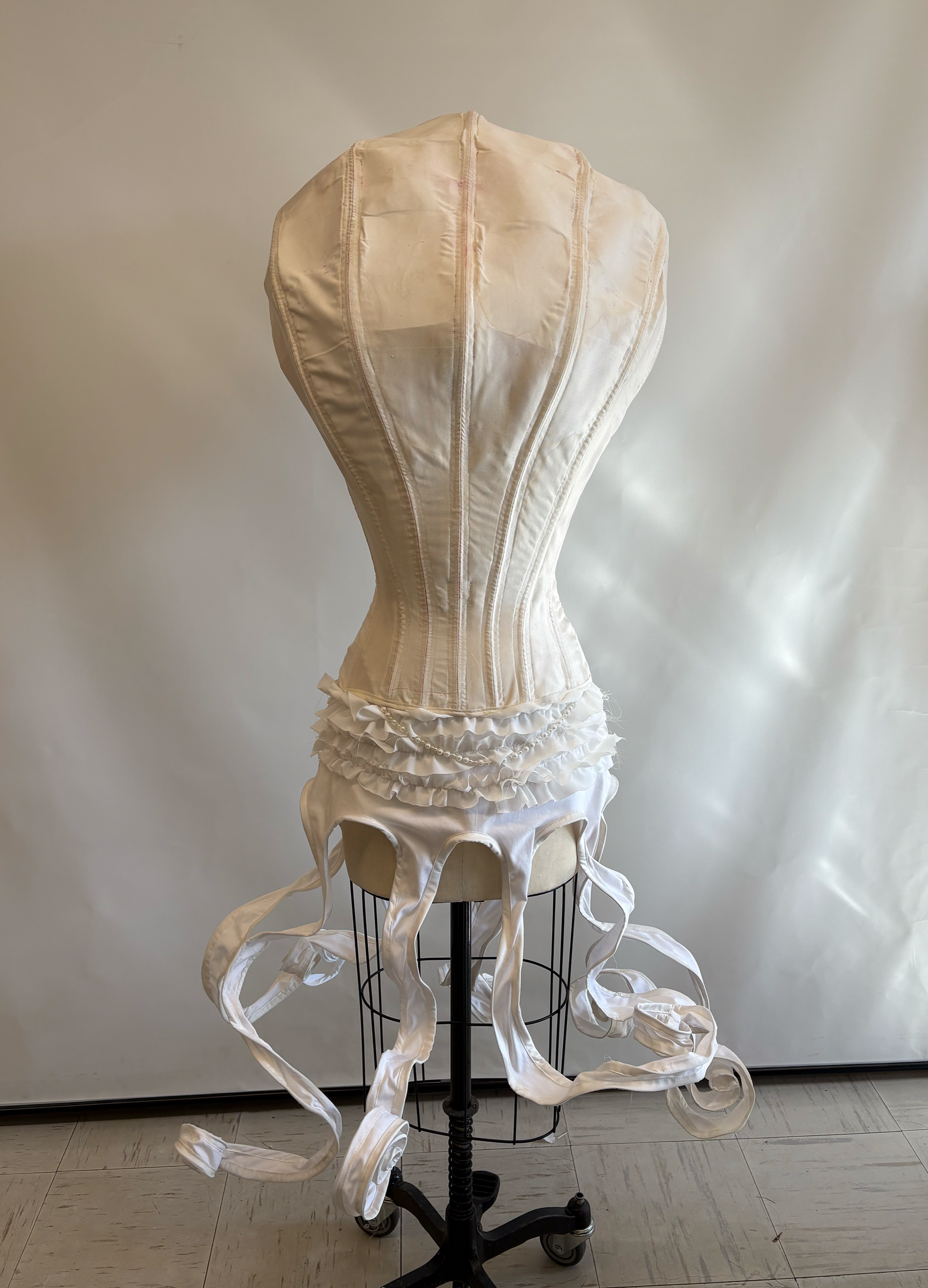 A vintage white dress form mannequin with a corset and a ruffled skirt, displayed on a black metal stand against a plain background.