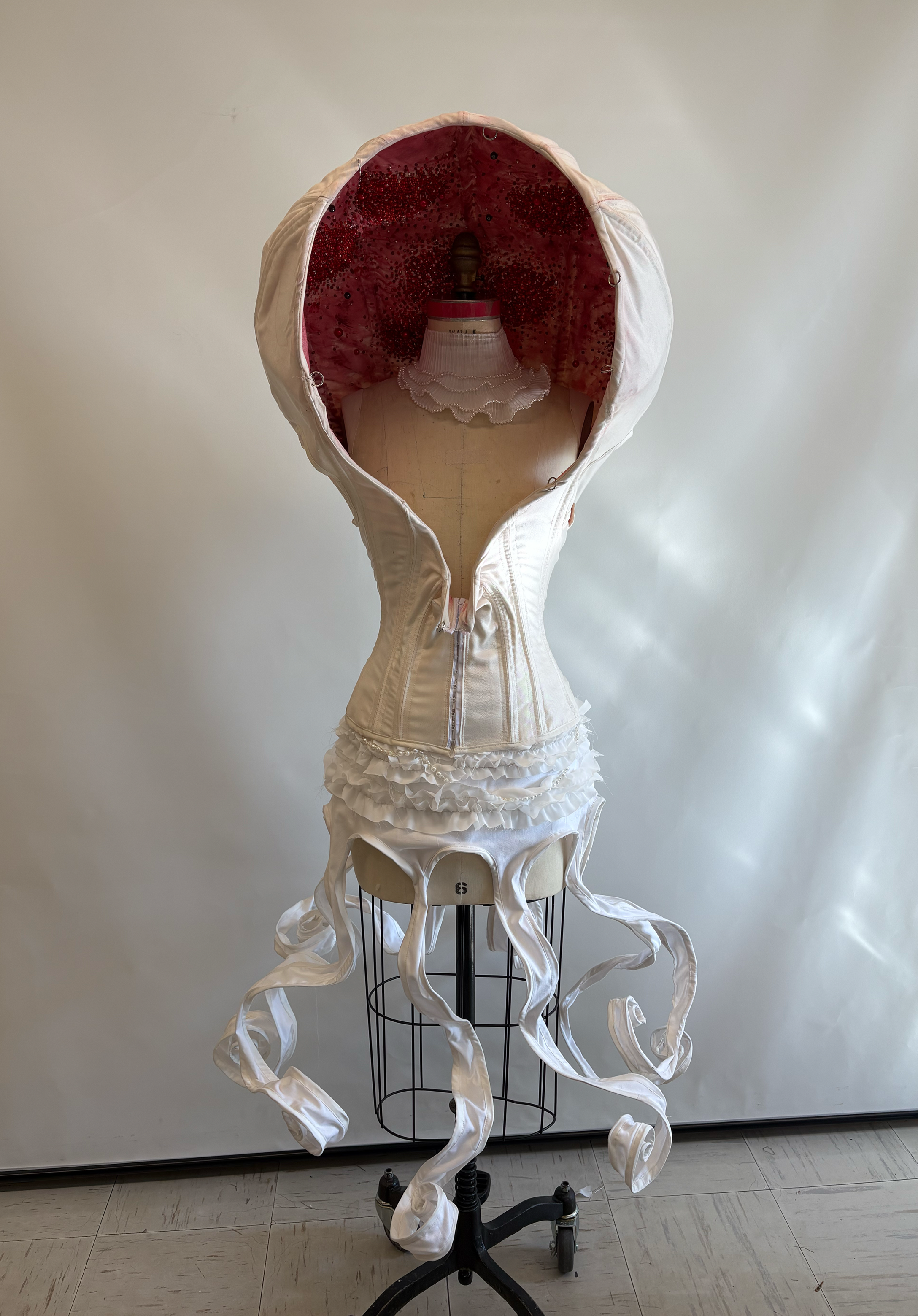 A dress form mannequin dressed in a white Victorian-style corset with ruffled layers and long, ribbon-like extensions, set against a plain wall.