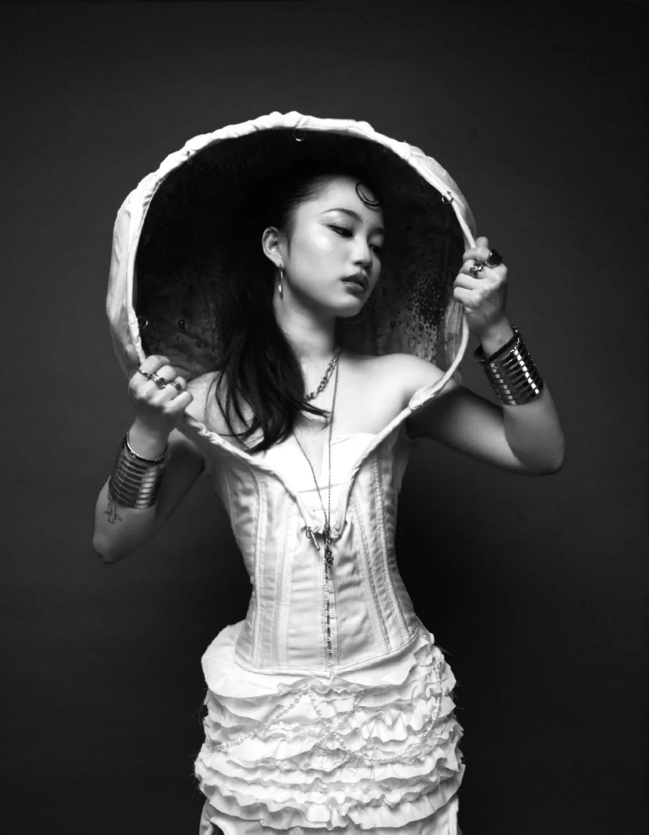 A woman wearing a corset and ruffled dress holds a large hooded umbrella. She has dark hair, earrings, and bracelets, and stands against a dark background, with a contemplative expression.