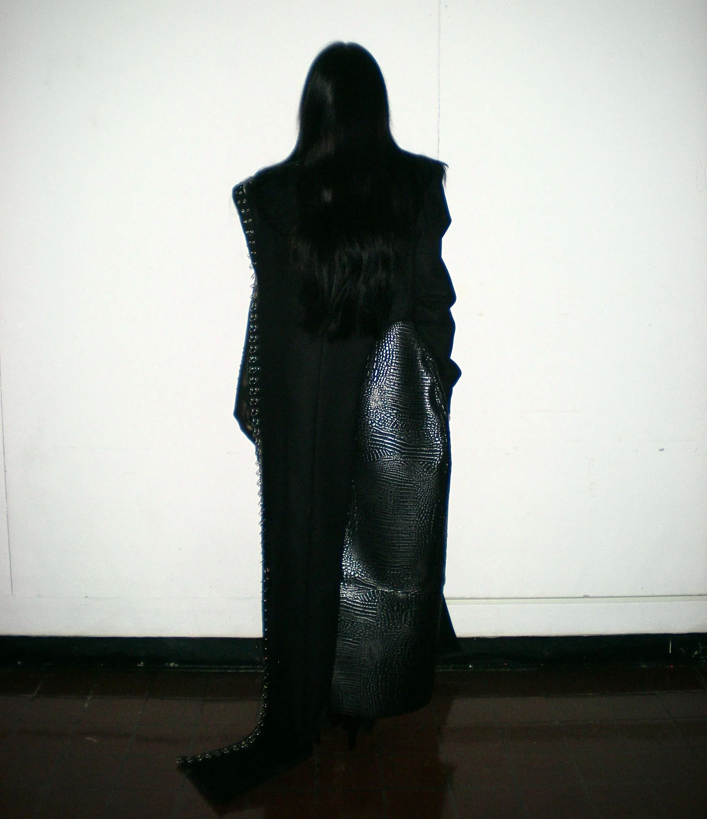 A person with long black hair wearing a black outfit with metallic details, standing against a white wall.