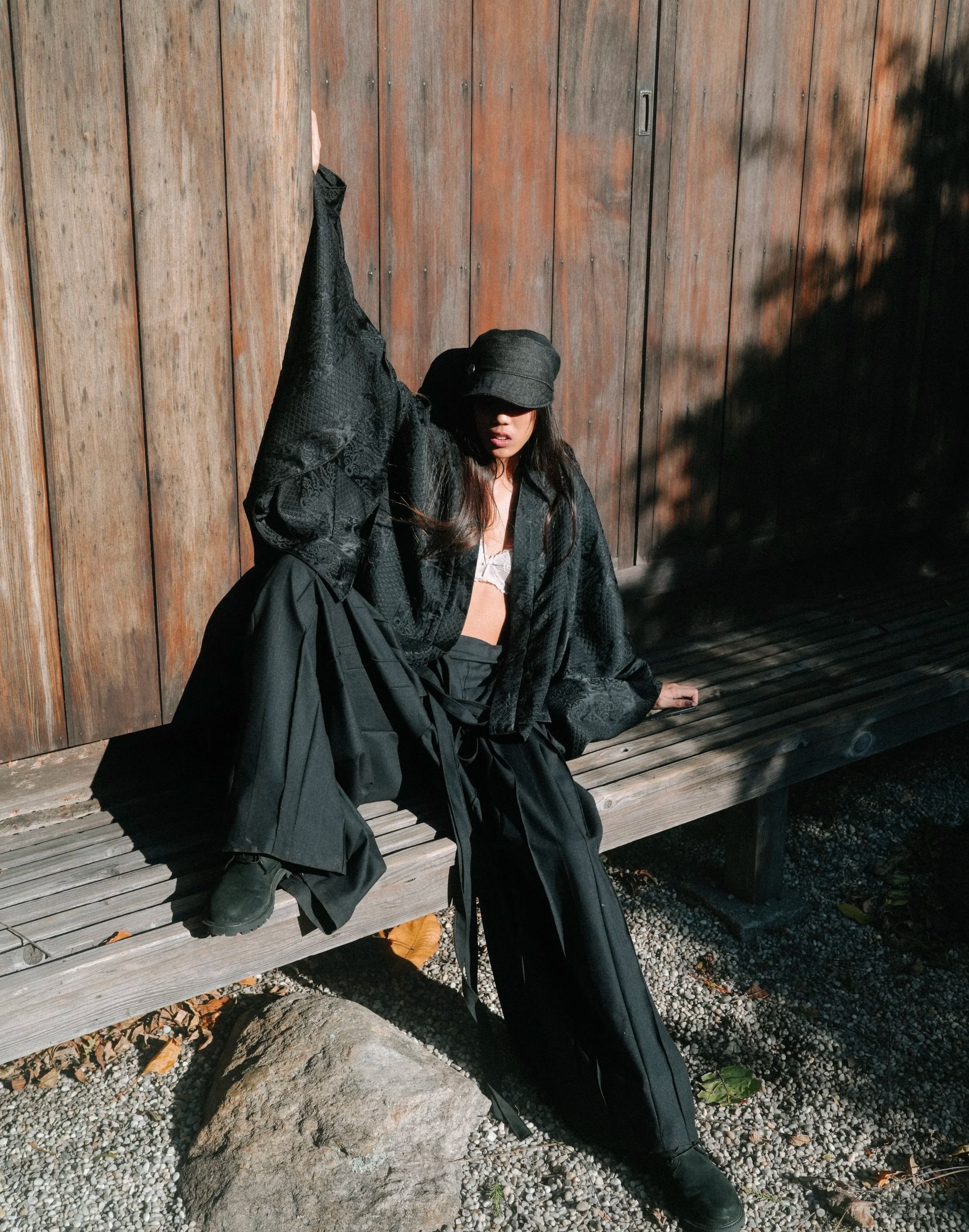 A woman wearing black clothing, a black hat, and a white bra sits on a wooden bench against a wooden wall, with shadows and sunlight on her and the background.