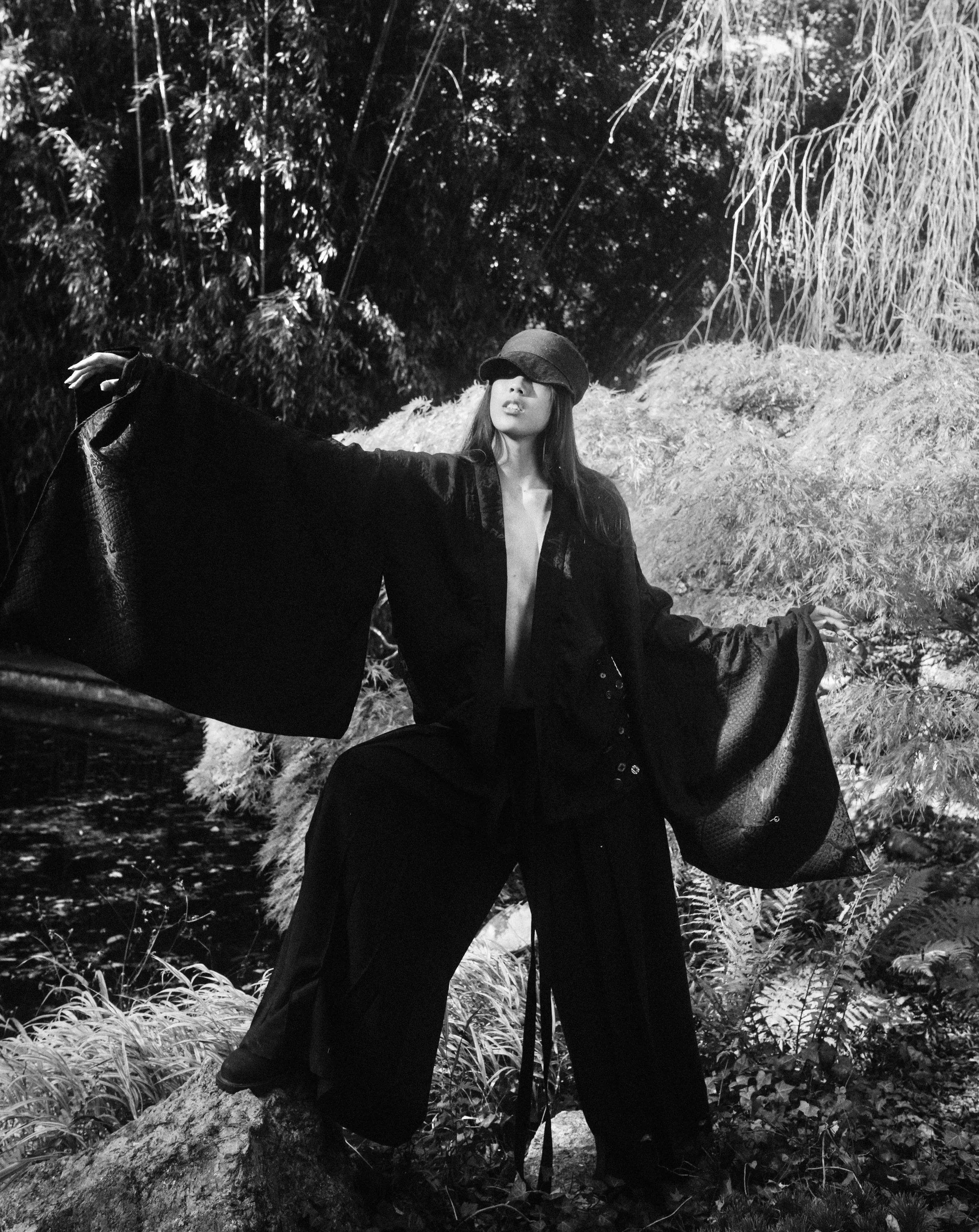 A woman with long hair is standing outdoors in a natural setting with trees and foliage, wearing a wide-brimmed hat and a loose, dark, flowing outfit, striking a pose with her arms extended.