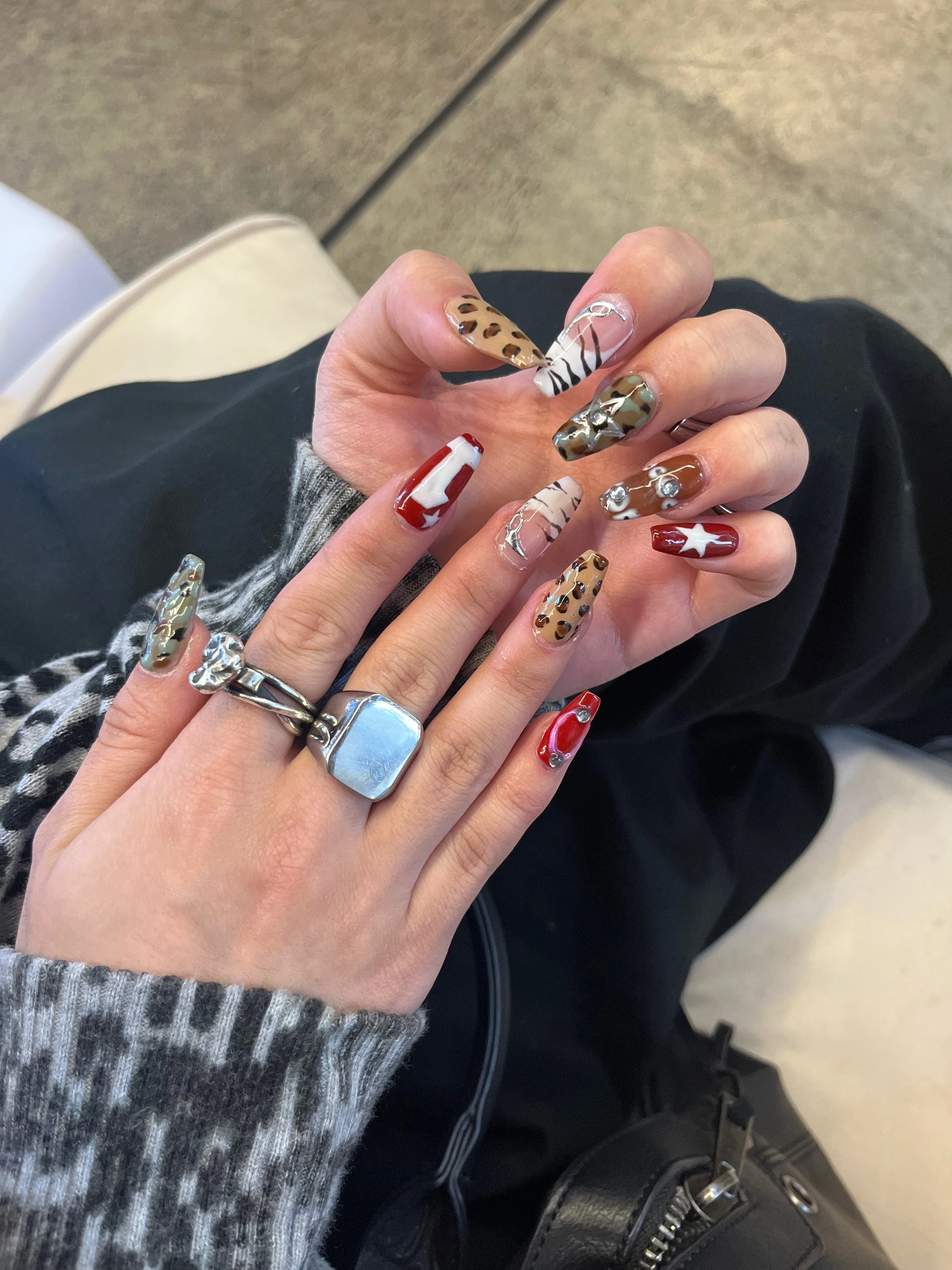 Close-up of a person's hand displaying decorated acrylic nails with animal print, red and white designs, and 3D embellishments, while wearing rings and a gray sweater.