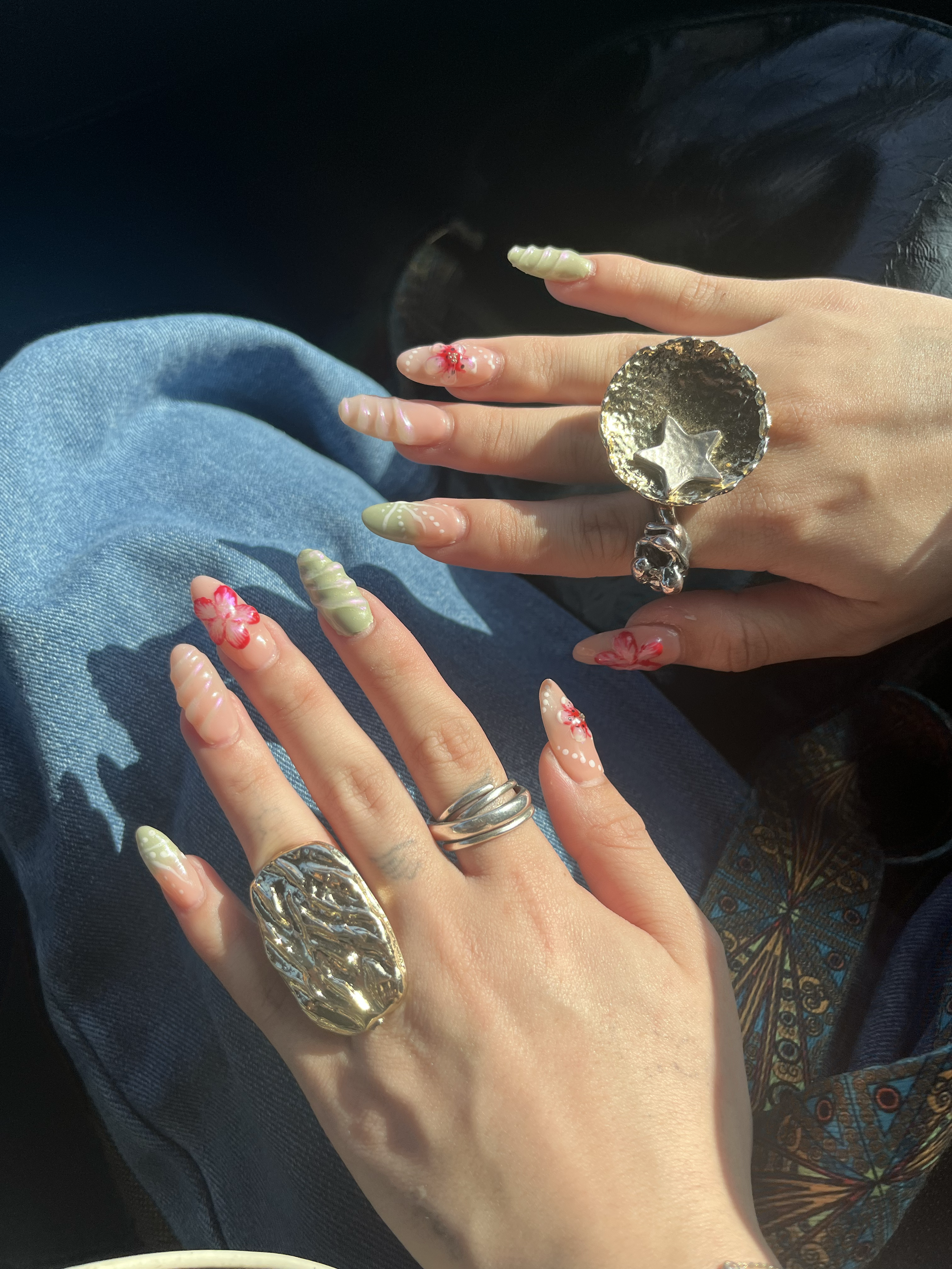 Close-up of two hands with ornate jewelry and decorated fingernails, resting on denim jeans. The nails are painted with pastel colors and floral designs.