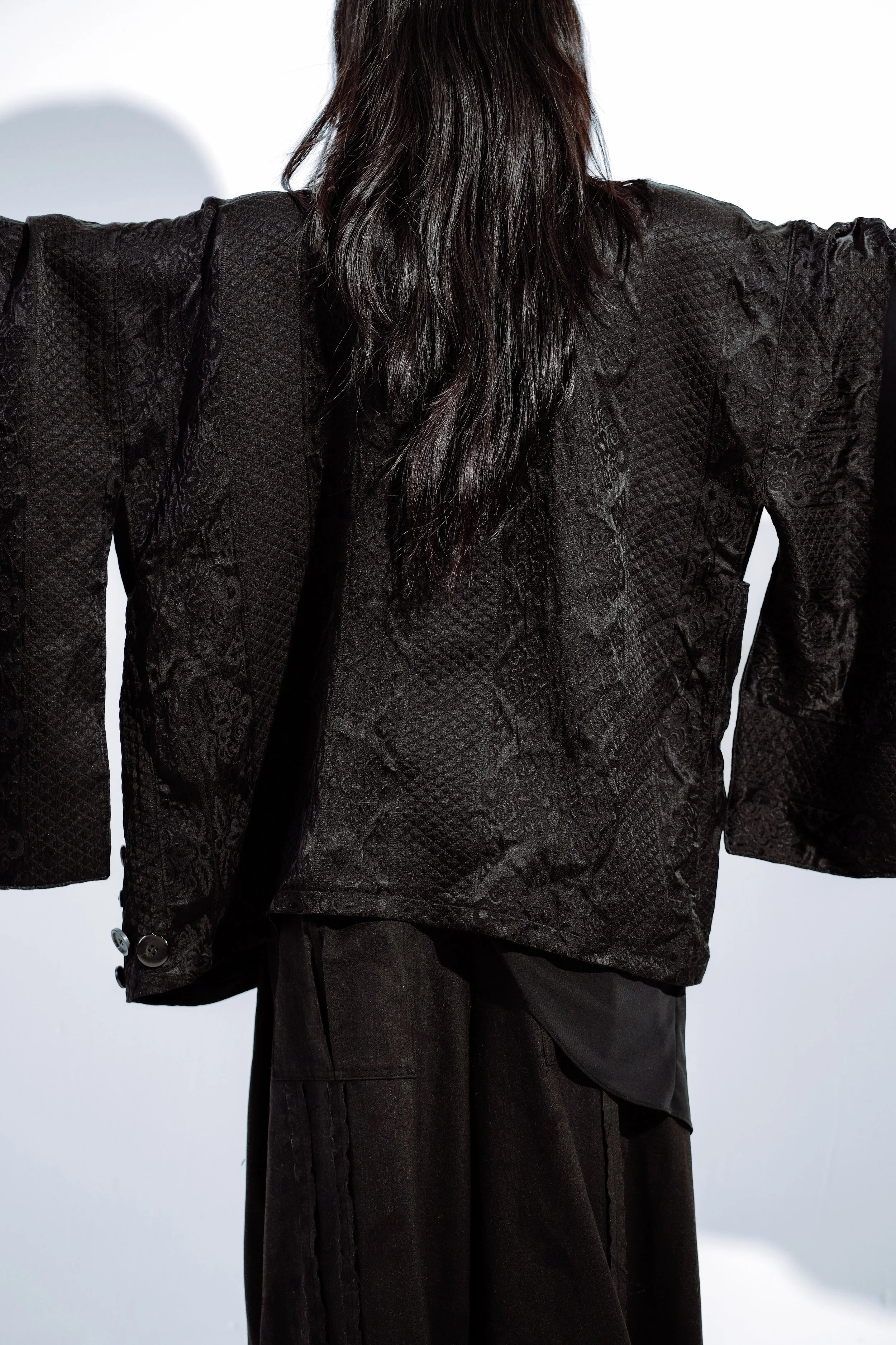 Person wearing a black, patterned blazer with wide sleeves, and black pants, standing against a white background.