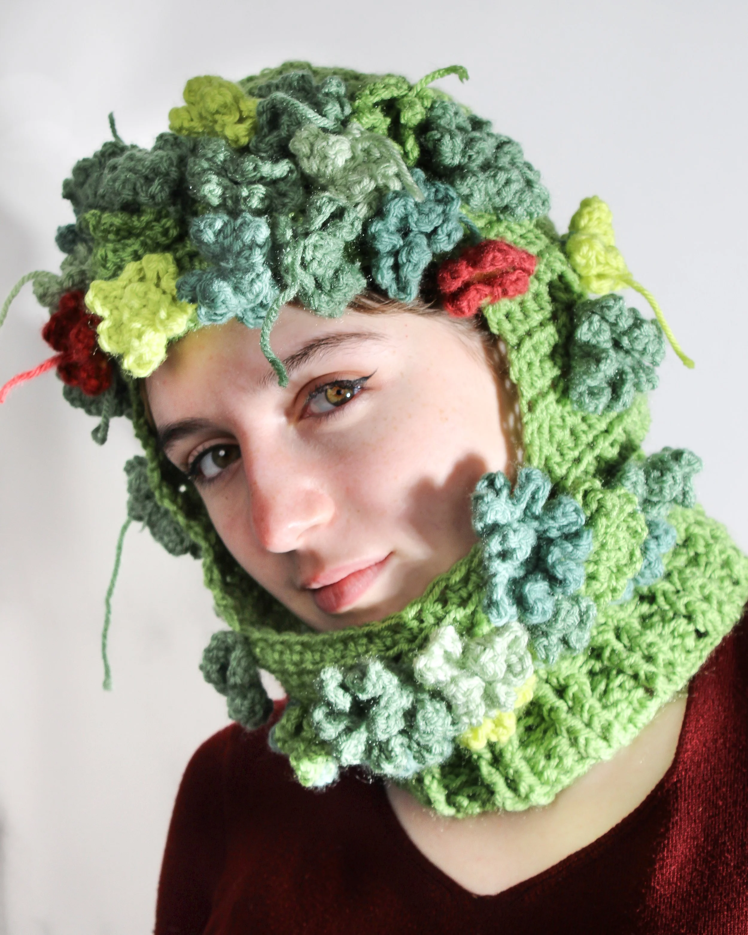 A woman wearing a hat and scarf made of colorful crocheted yarn designed to look like green plants and red flowers.
