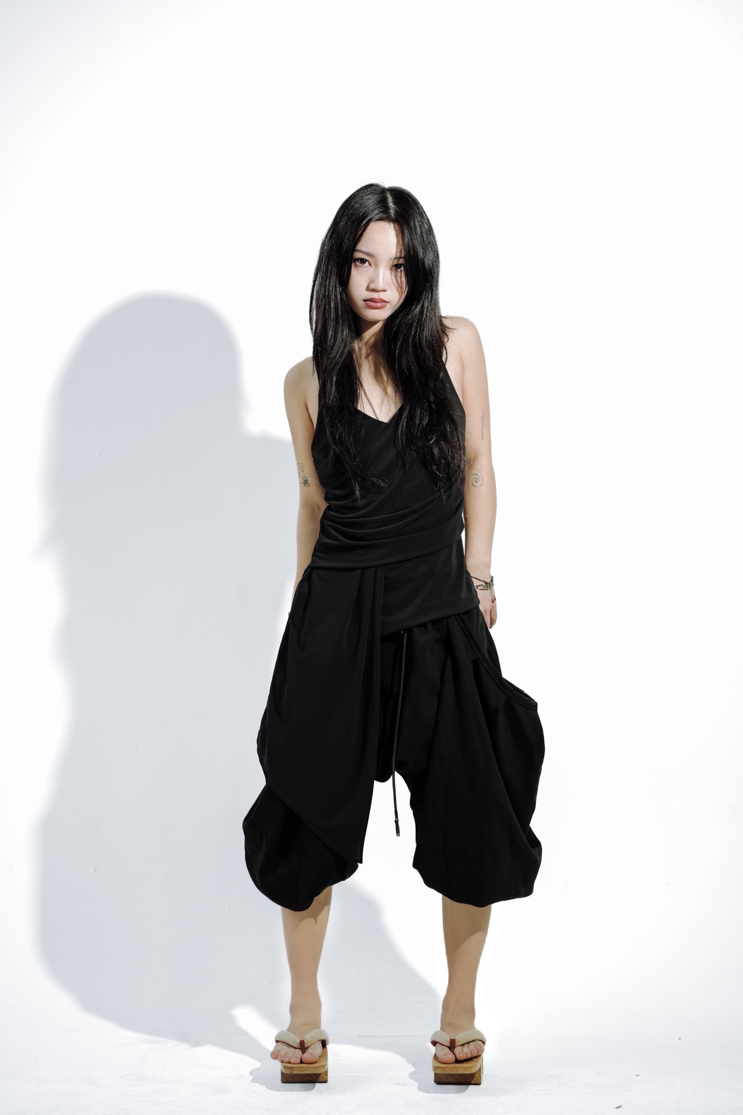 A young woman with long black hair wearing a black sleeveless top and baggy black pants, posing against a white background with her shadow cast on the wall.