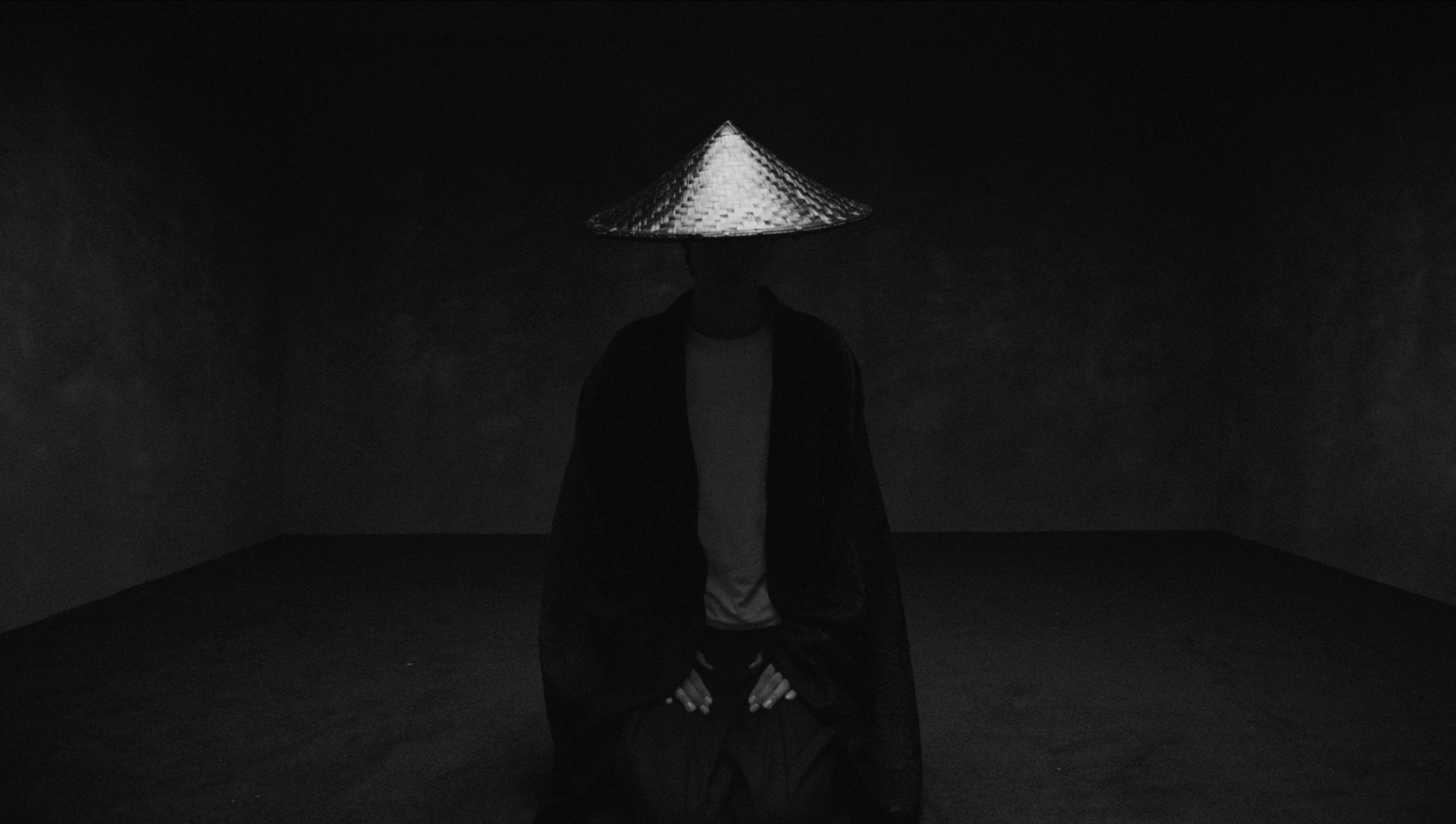 Person sitting cross-legged with hands on knees, wearing a conical Asian hat, in a dark, minimal room.