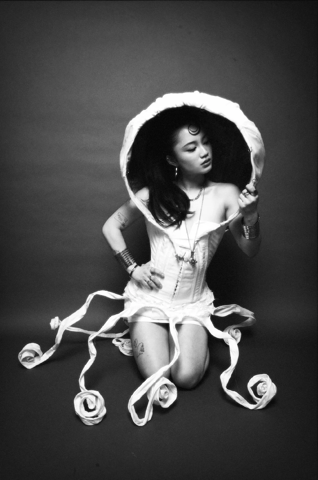 A black and white photo of a woman kneeling on the ground, wearing a corset and a large hooded piece with fabric strips resembling tentacles.