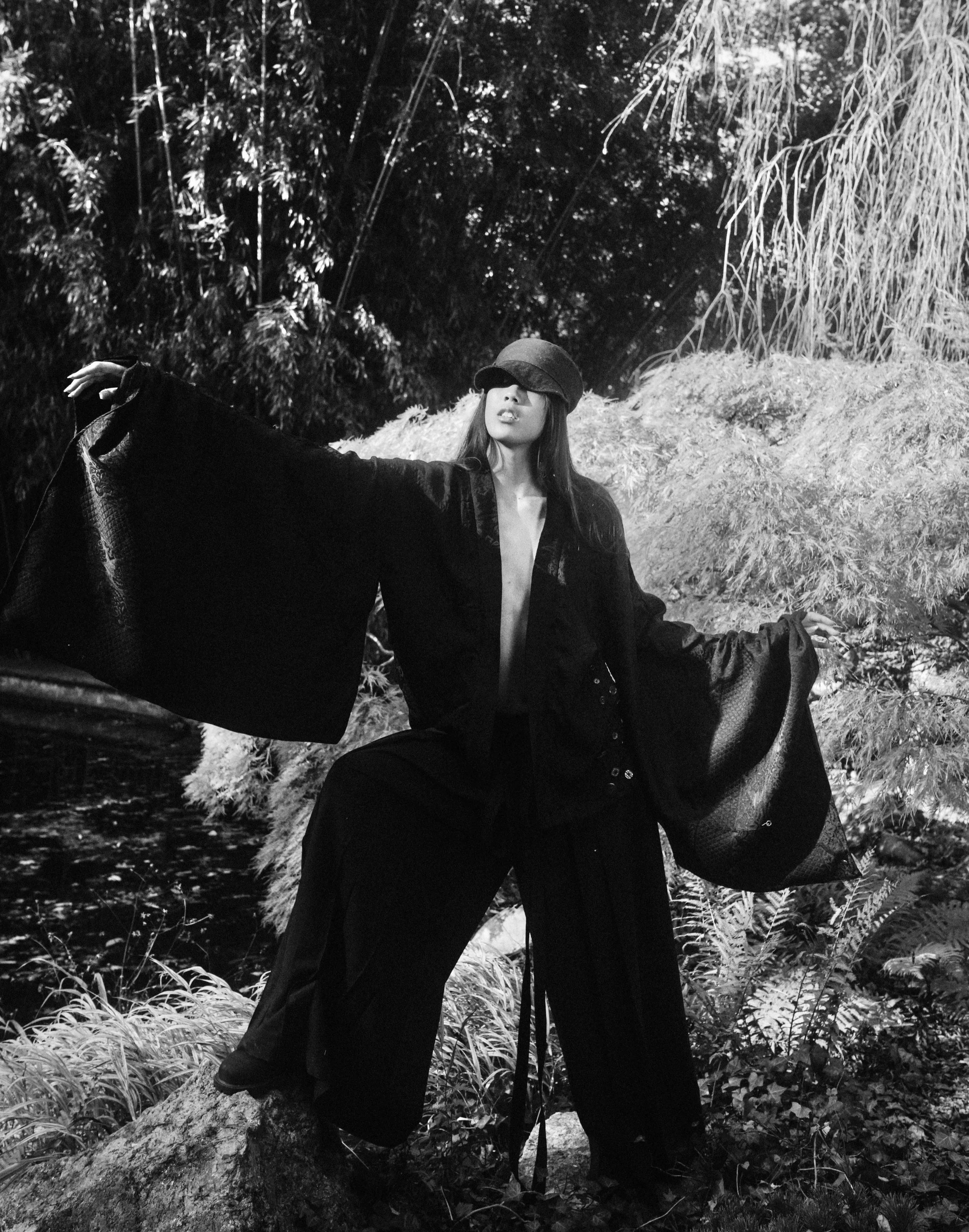 A woman wearing a hat and a long, flowing black coat standing in a natural outdoor setting with trees and foliage, striking a pose with arms outstretched.