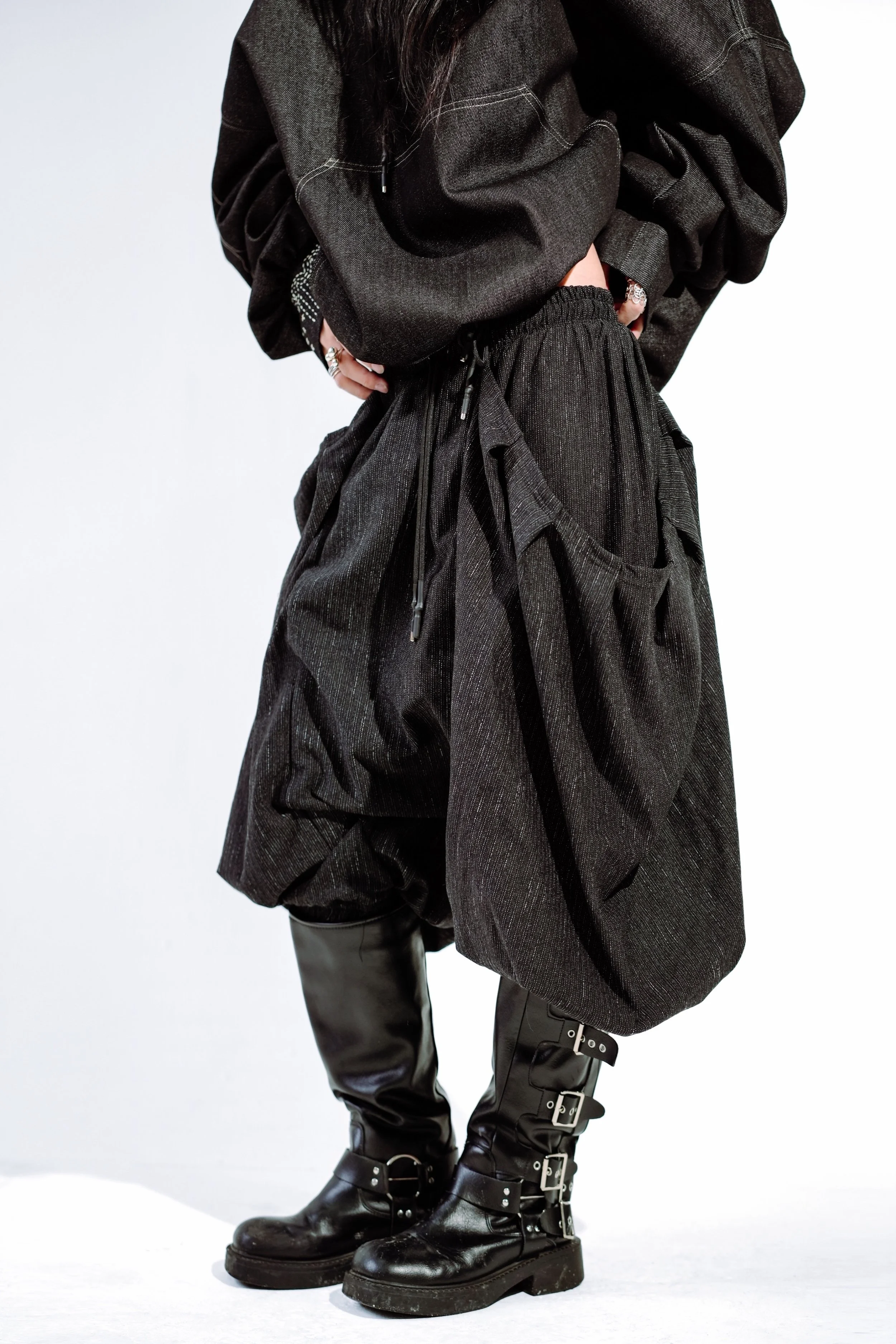 Person wearing black clothing, including baggy pants and boots with buckle details, standing against a plain background.