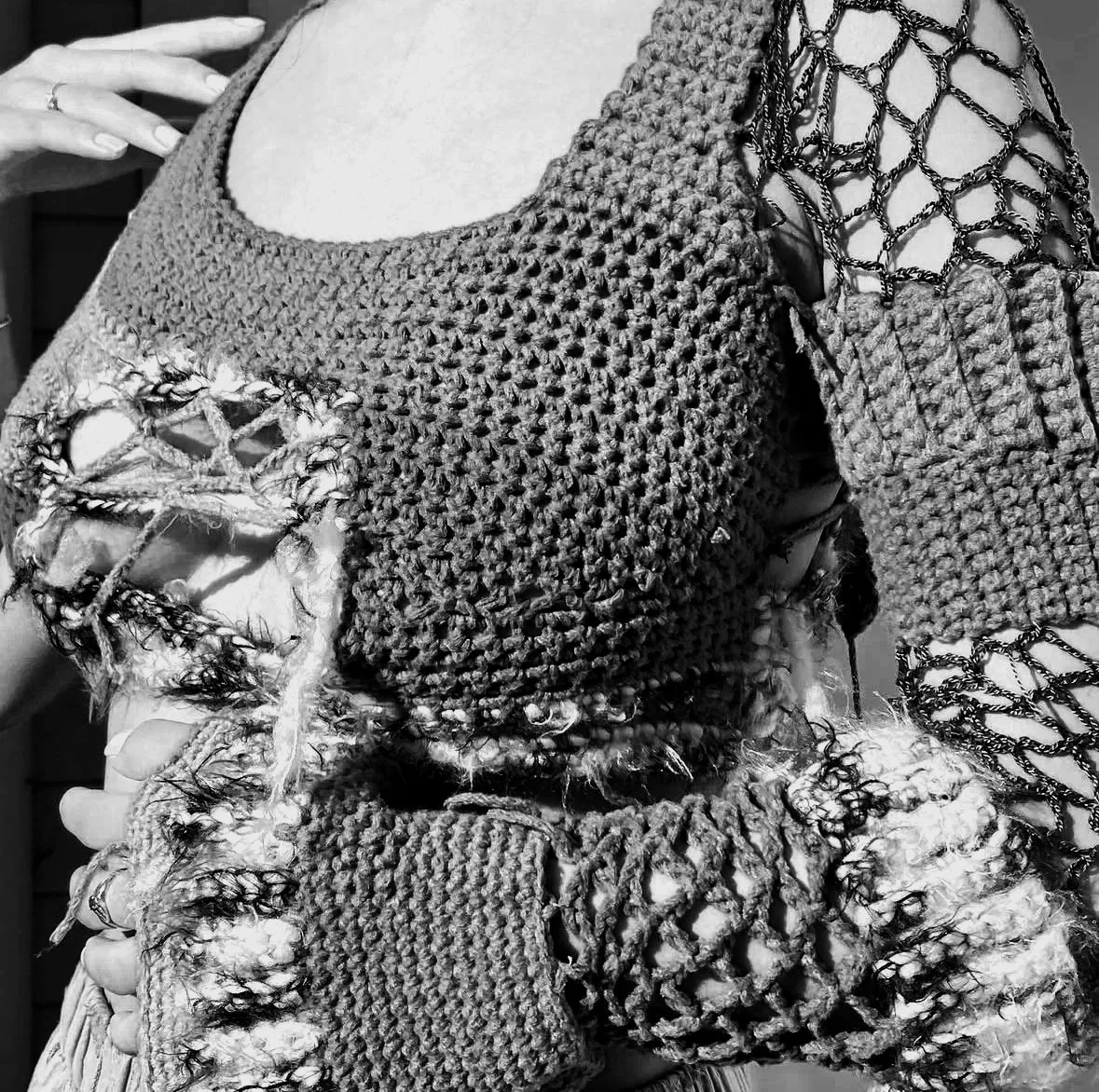 Close-up of a person wearing a textured, distressed knitted sweater with open weave details and decorative chain embellishments on the shoulder and sleeve, captured in black and white.