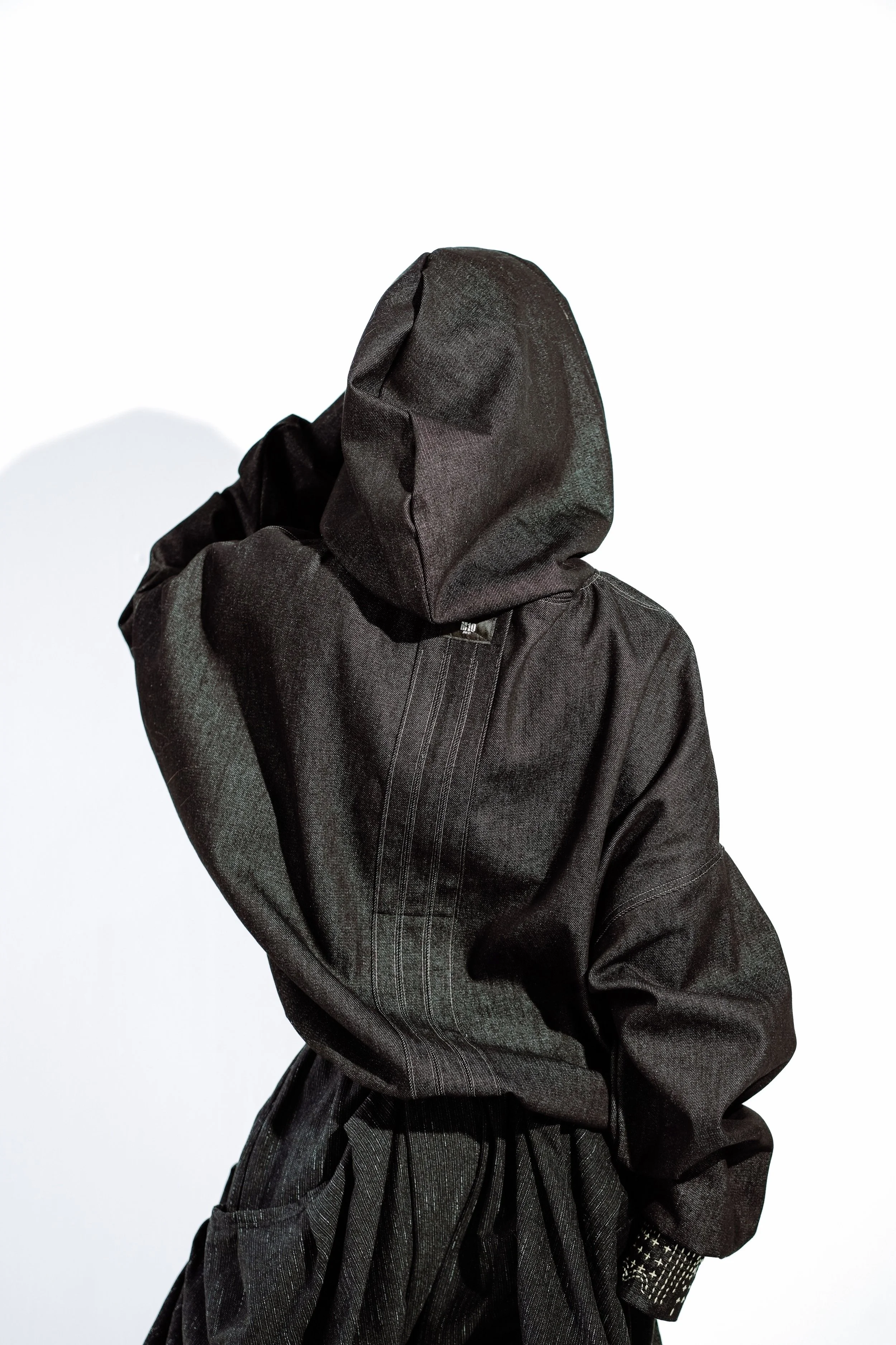 Person wearing a black hooded jacket with the hood up, touching their head, against a white background.