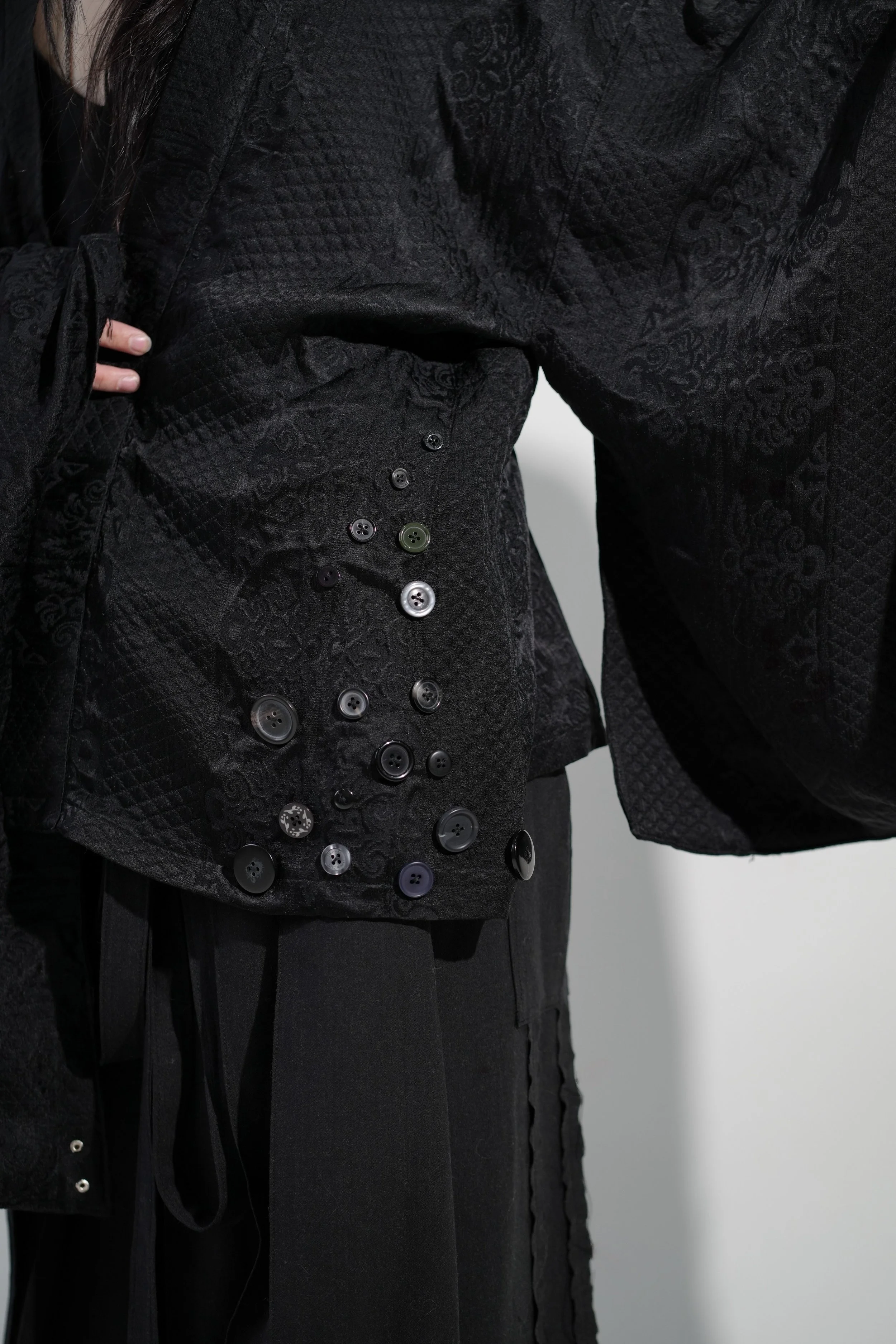 Close-up of a black quilted jacket with various black, grey, and green buttons on the sleeve, worn over black pants.