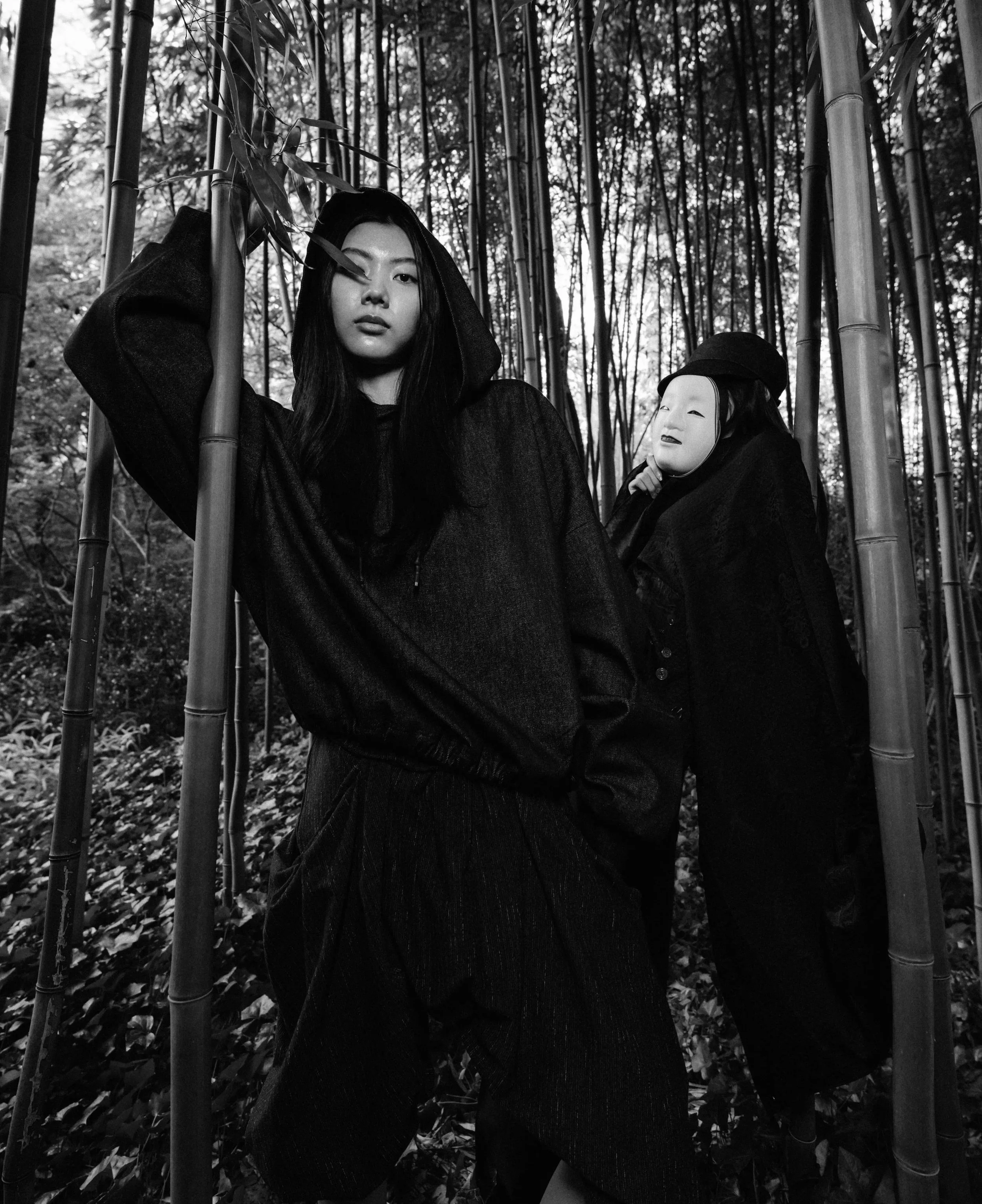 A woman dressed in dark clothing standing in a bamboo forest holding a bamboo stalk, with a mask of a smiling face hanging on a stick behind her.