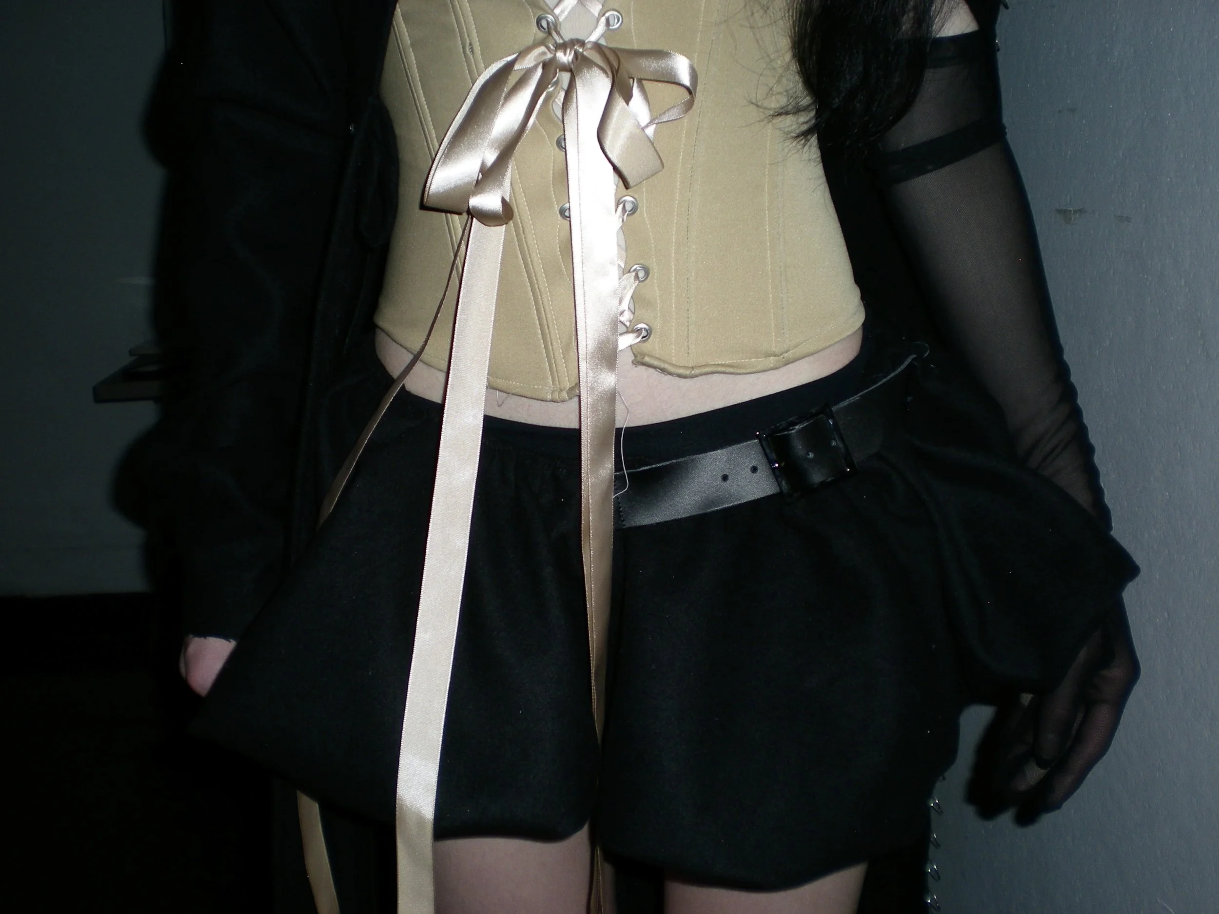 Close-up of a person wearing a beige corset with a satin ribbon, black shorts with a belt, and black mesh long sleeves, holding a black bag with beige ribbons.