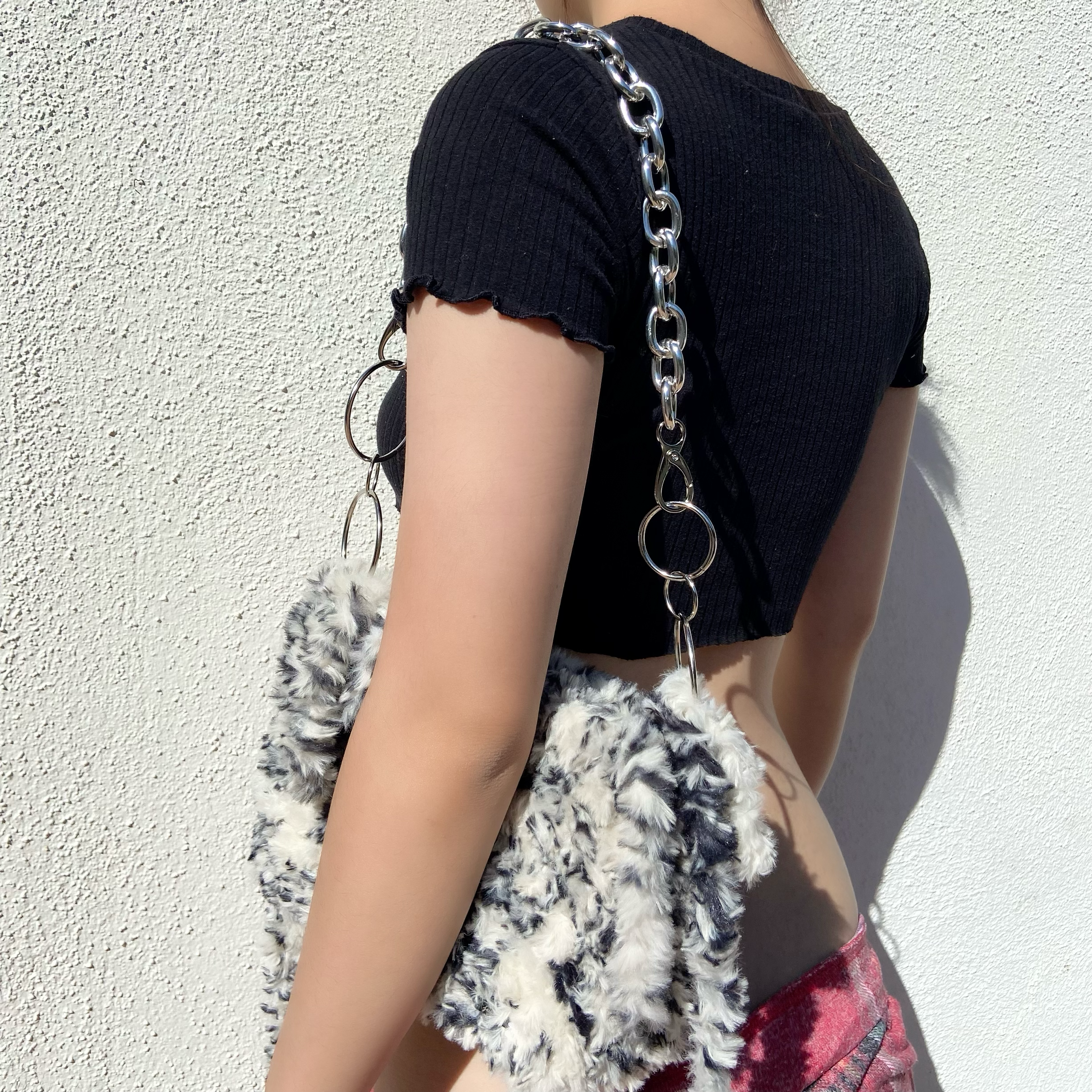 Person wearing a black crop top with ruffled sleeves, carrying a fluffy patterned bag with a silver chain strap, standing against a white textured wall.