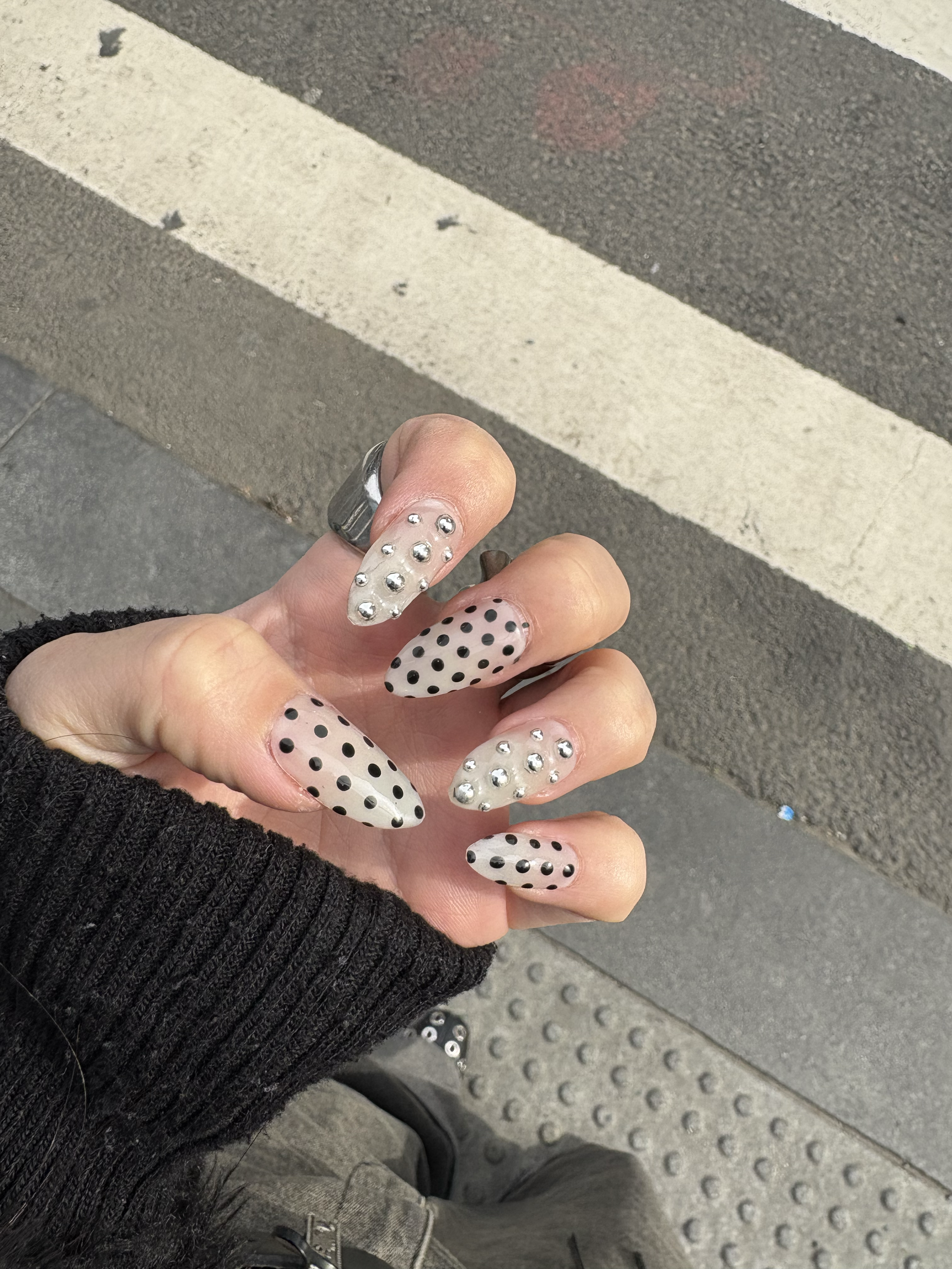 Hand with decorated nails featuring black polka dots on a beige background, some nails with silver studs; background shows a street curb and pavement.