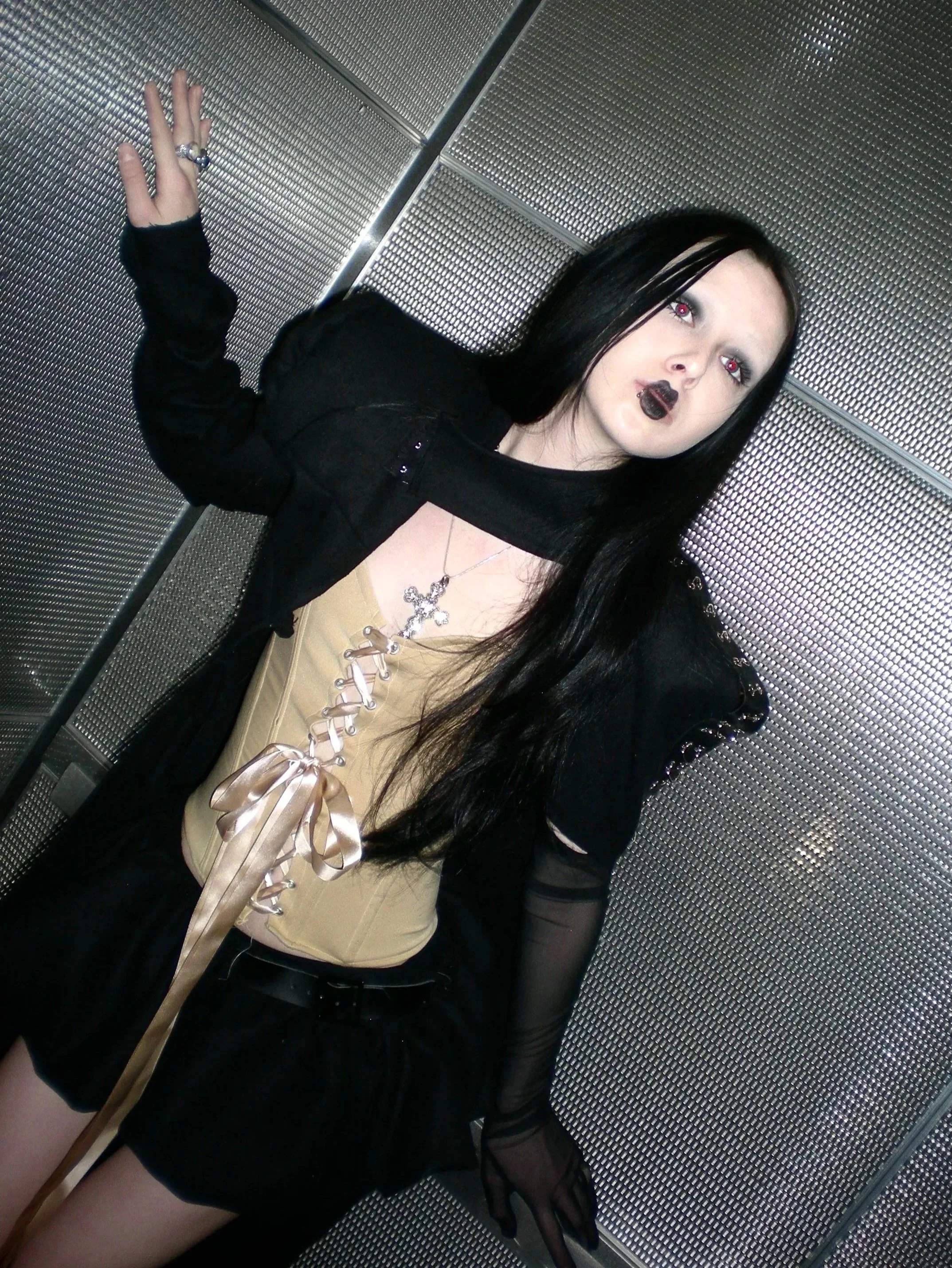 A woman with long black hair and Gothic makeup, wearing a black jacket, a beige corset with ribbon lacing, and a gold cross necklace, posing in an elevator with metallic walls.