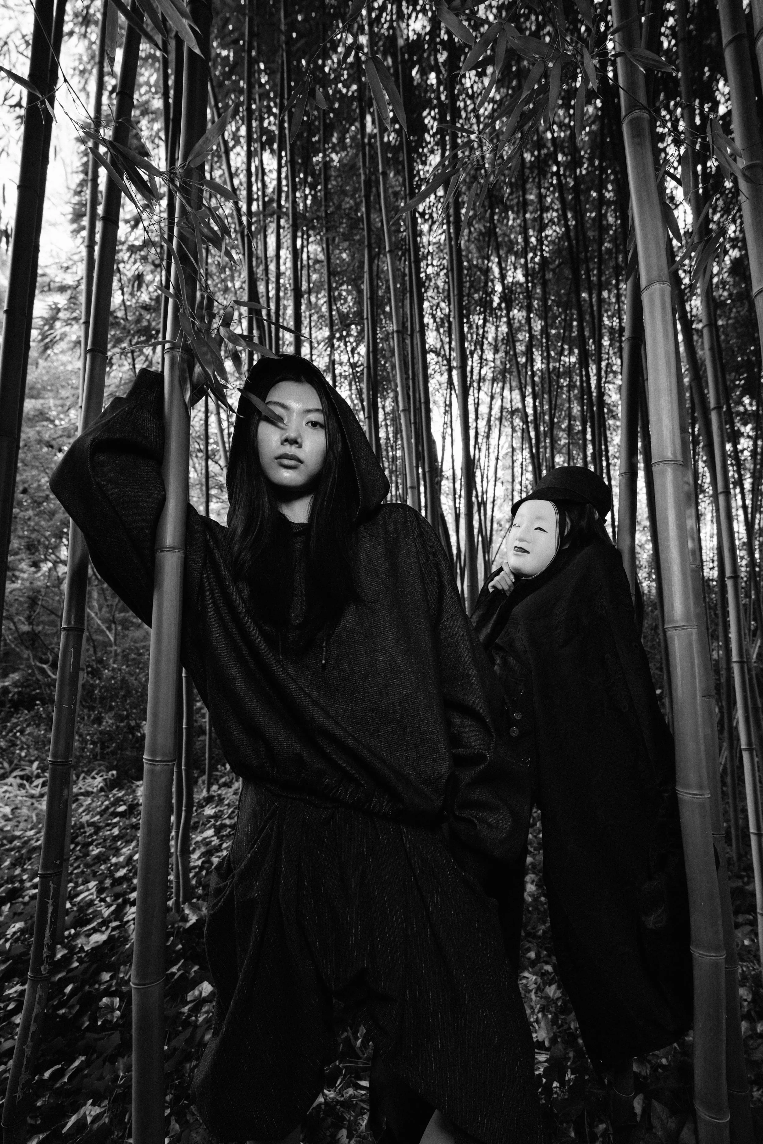 A black and white photo of two women in a bamboo forest, one with long dark hair wearing a hoodie, and the other with a porcelain mask and traditional clothing.