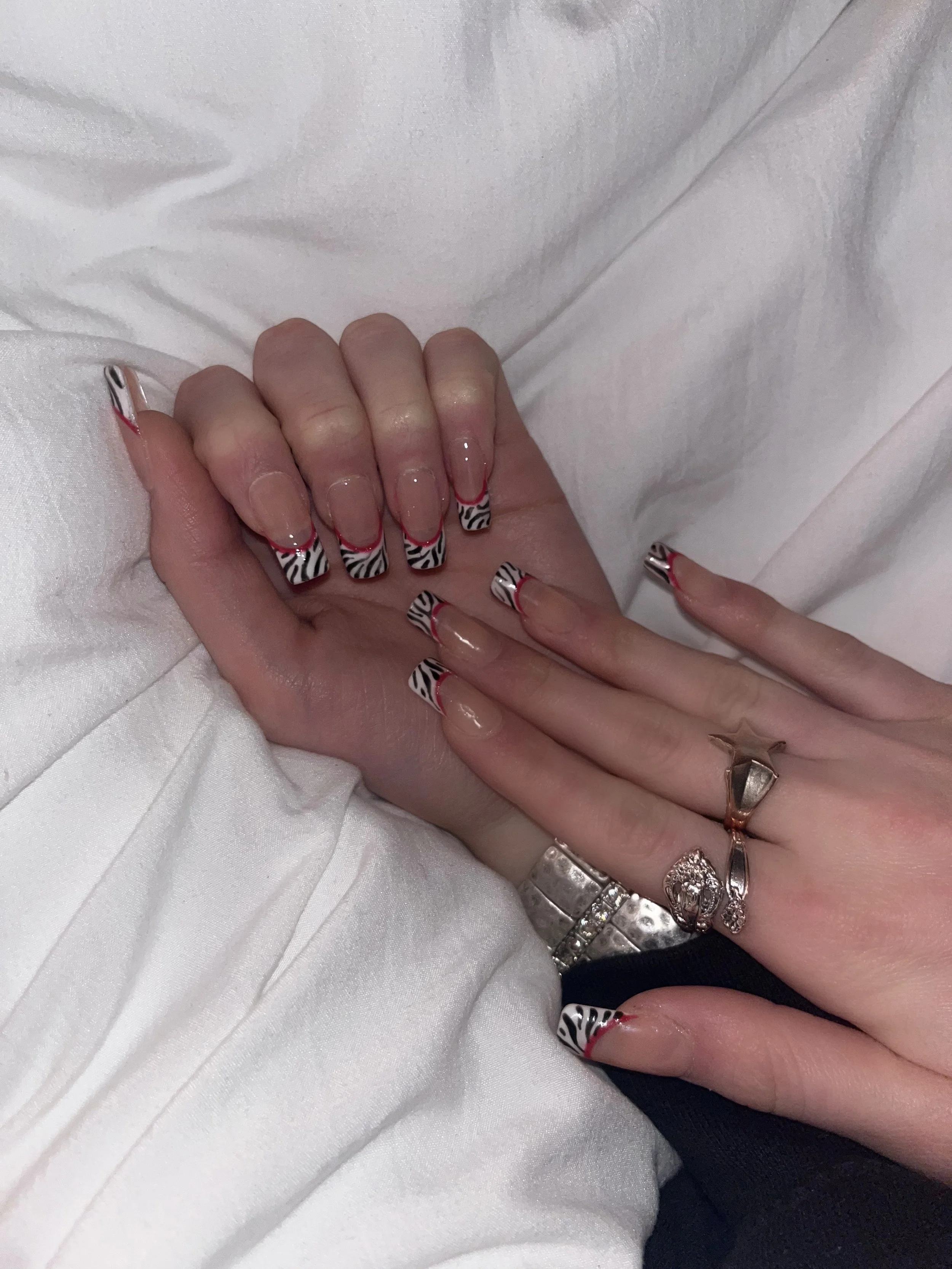 Close-up of two hands with decorated fingernails, featuring zebra stripe nail art with red tips and a clear base, resting on a white fabric surface. The hands are adorned with rings, including a silver band and rings with stone embellishments.
