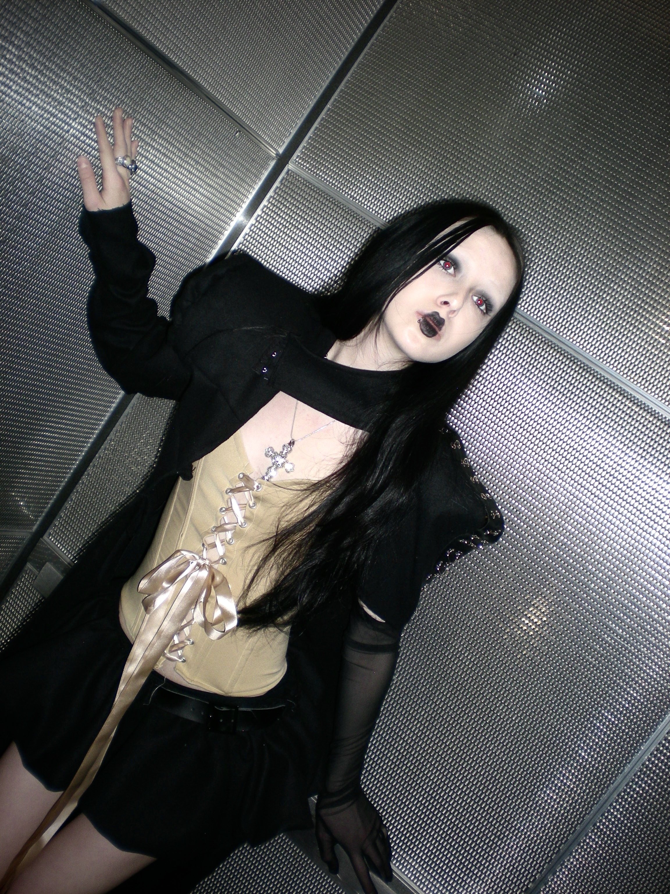 A young woman with long black hair and gothic style makeup, wearing a beige corset with a ribbon front, black blazer with decorative studs, black skirt, and sheer black gloves, standing inside an elevator with metallic walls.