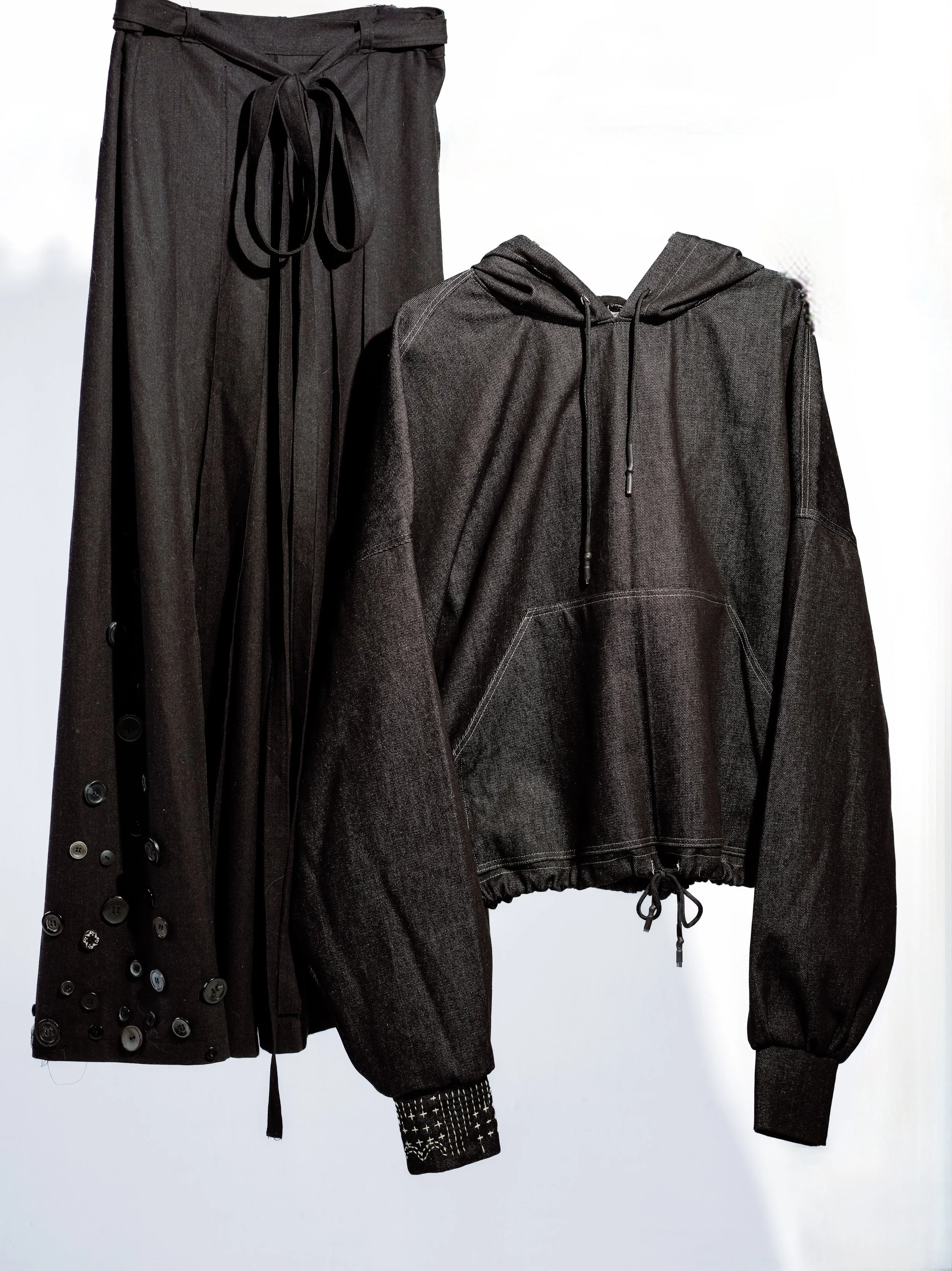 A black pair of pants with a drawstring waist, decorated with buttons on the side, hanging next to a black hooded jacket with a zipper and drawstrings.