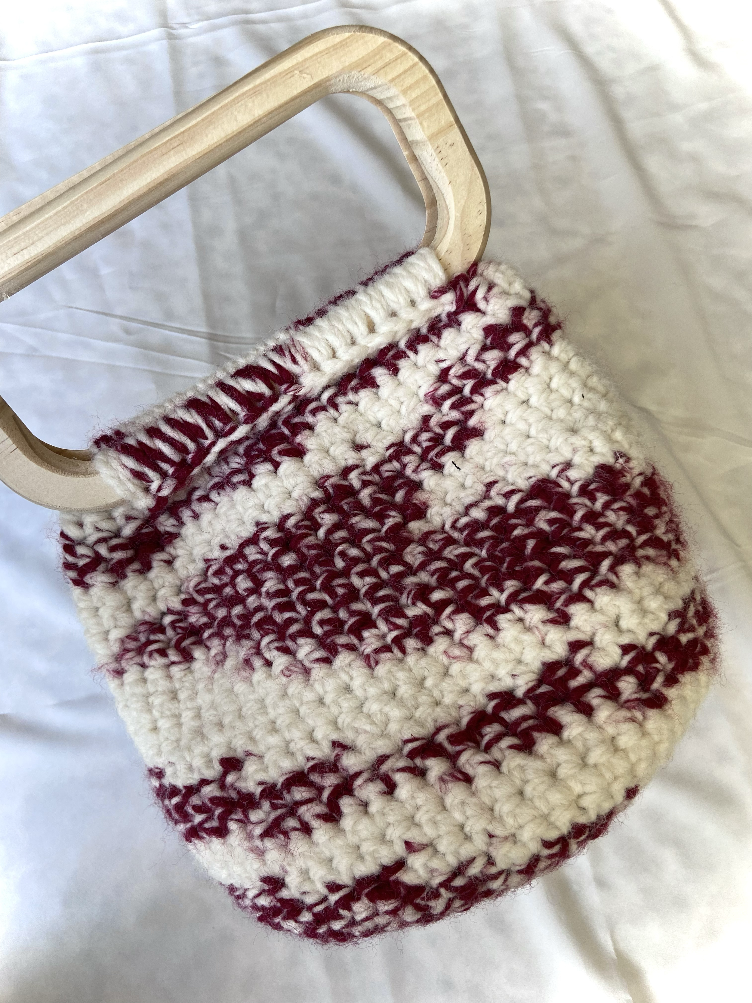 A knitting project in progress on a wooden needle, made with white and maroon yarn in stripes, resting on a white fabric background.