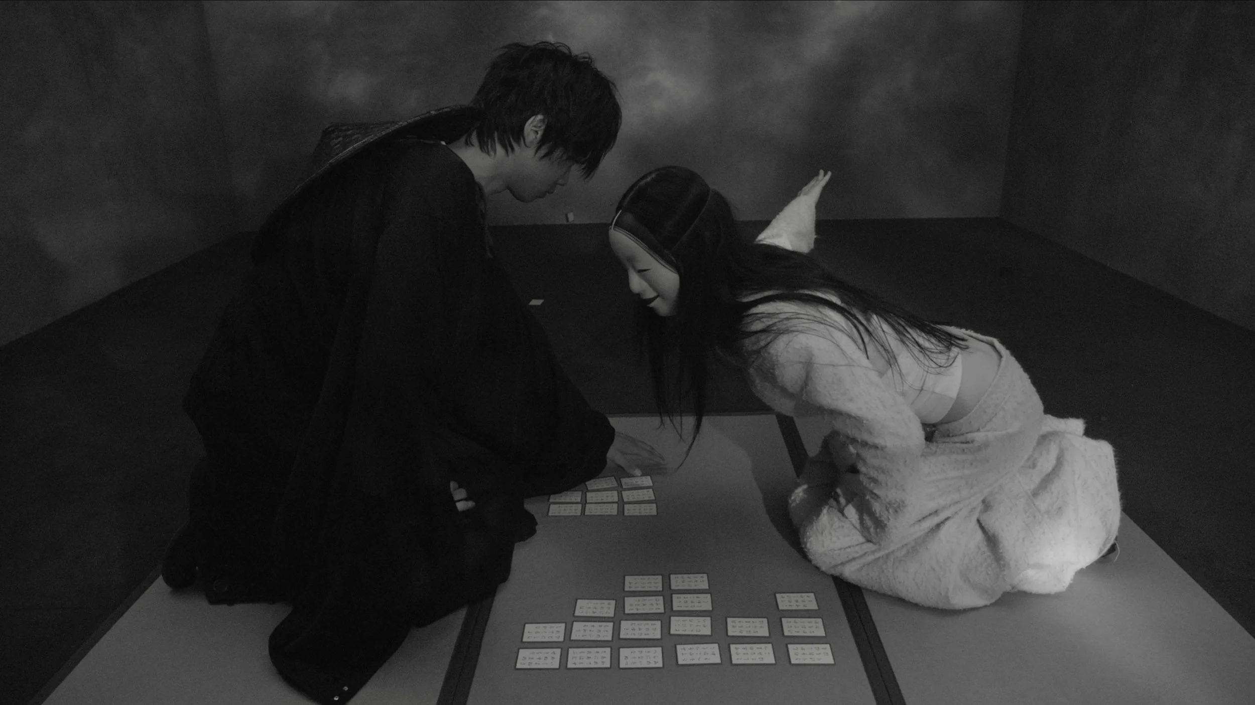 Two people wearing headphones are kneeling on the floor, playing a card game on a low table in a dimly lit room with dark walls.