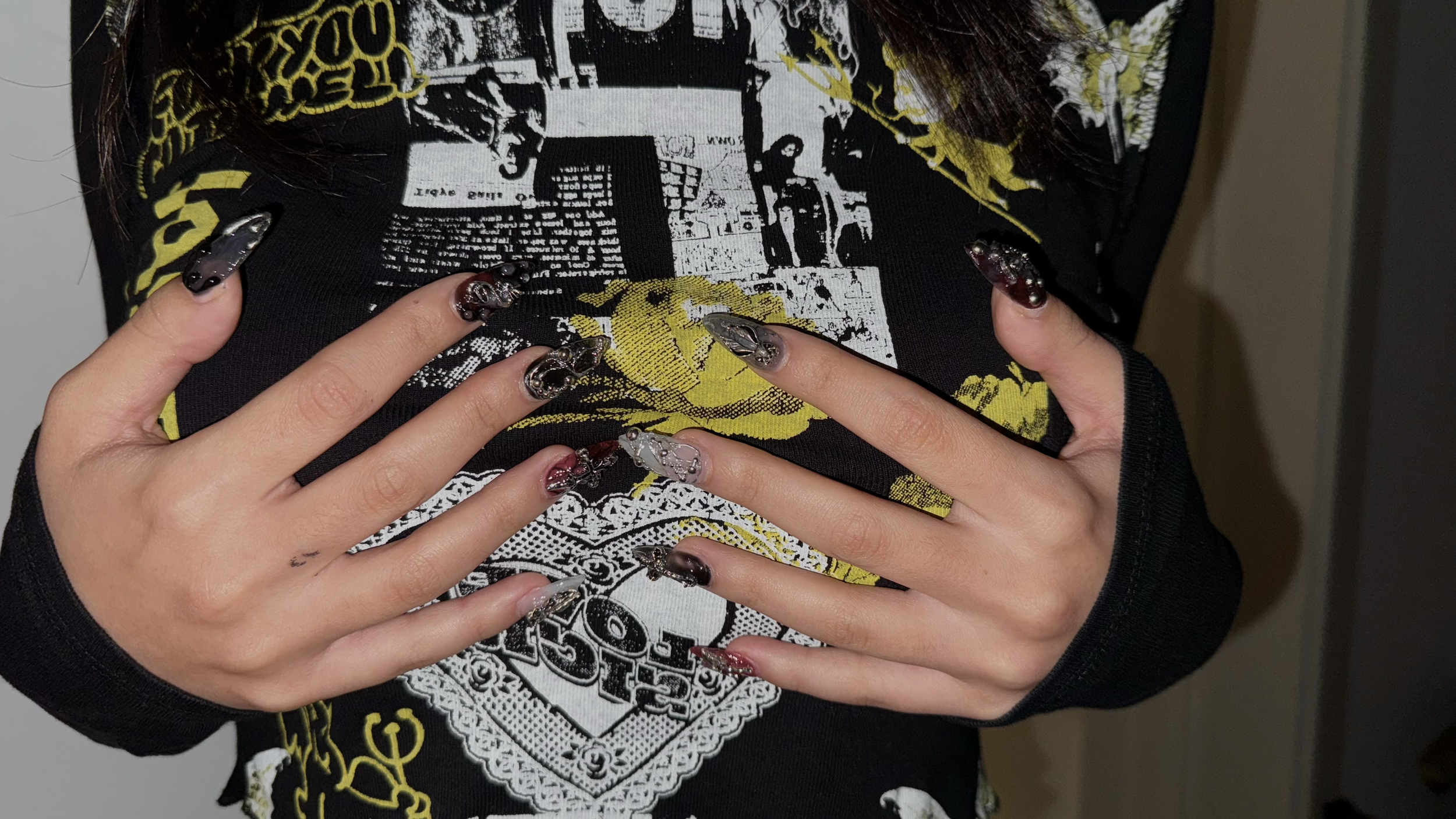 Close-up of a person with decorated nails, wearing a black graphic sweatshirt, touching their chest with both hands.