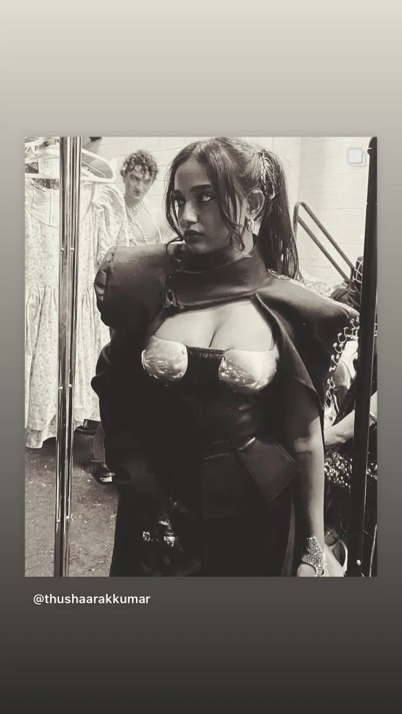 A woman with dark hair in pigtails wearing a shiny, futuristic-style outfit, standing in front of a mirror with a person in a patterned dress in the background.