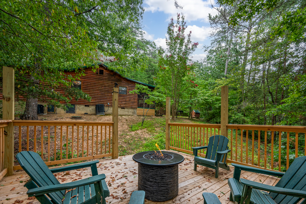 A wooden deck with four green Adirondack chairs and a fire pit in the center, surrounded by trees and a house in the background.