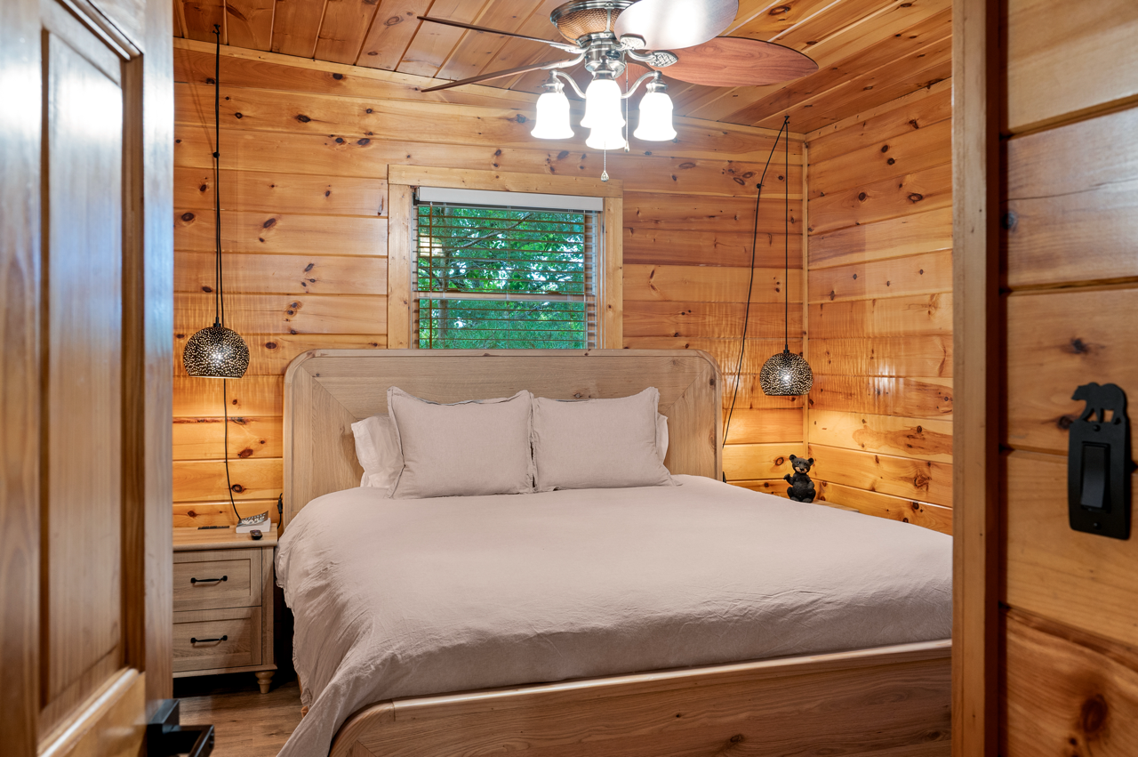 A cozy bedroom with wooden panel walls, a wooden bed frame with beige bedding and pillows, two hanging pendant lights on either side, a small nightstand with a lamp and remote, a window showing green trees outside, and a ceiling fan with lights.