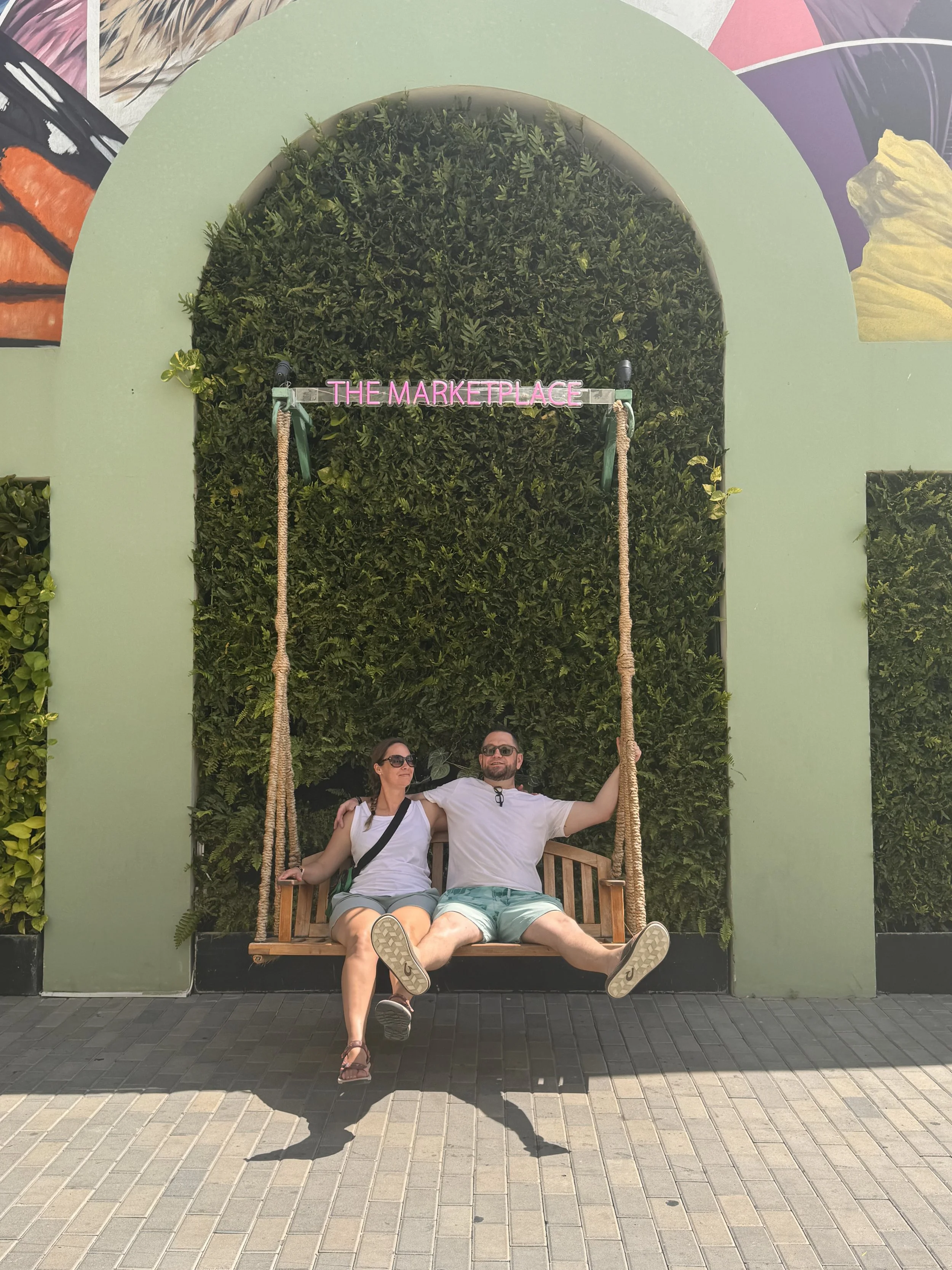 A man and woman sitting on a wooden swing with a green wall and hedge background, and a pink neon sign reading 'The Marketplace' above them.