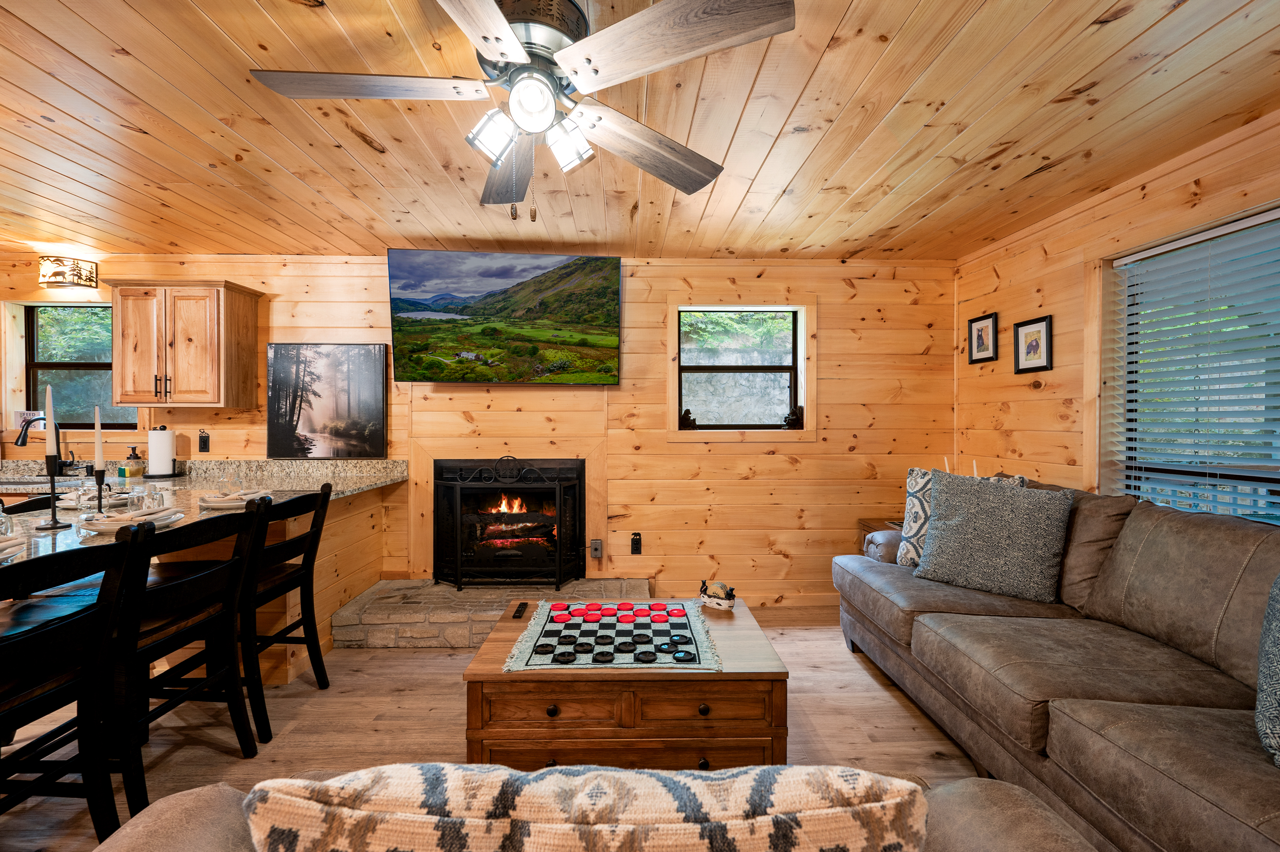 A cozy living room in a log cabin with wood-paneled walls and ceiling, featuring a fireplace, a wall-mounted TV, a brown sofa with pillows, a coffee table with a checkers game, and a window with blinds.