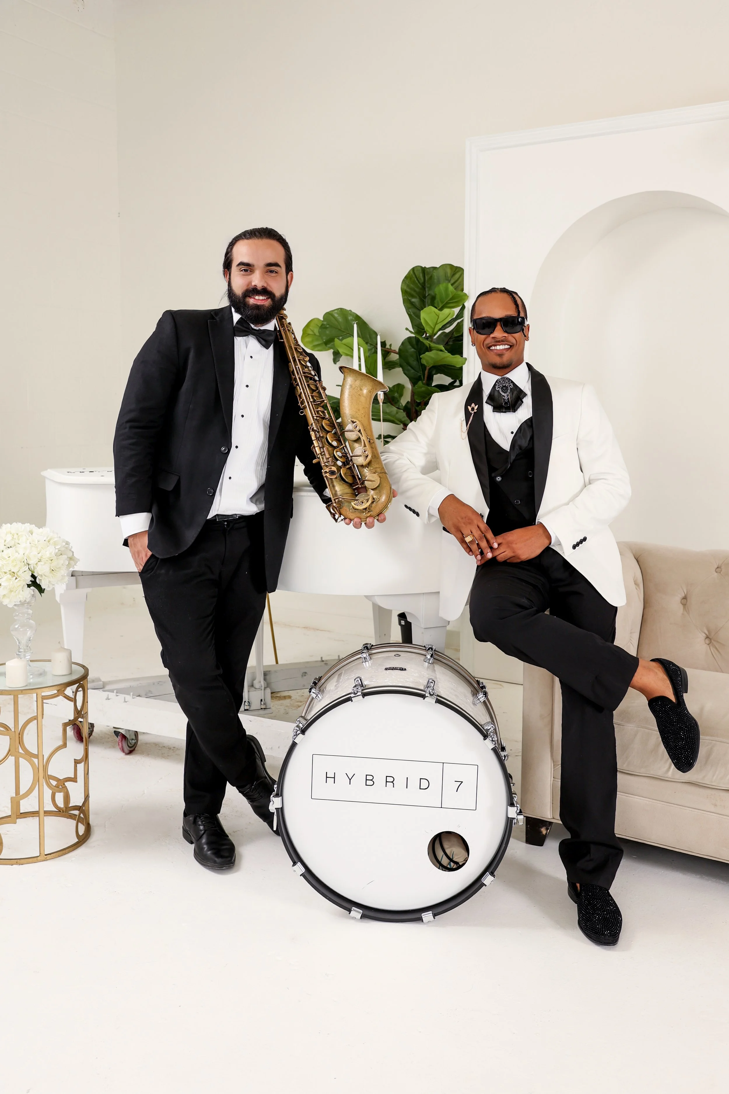 Two men in tuxedos with musical instruments, one holding a saxophone and the other wearing sunglasses, standing near a white piano and a large white drum with the text "HYBRID 7," in a room with white walls and a beige sofa.