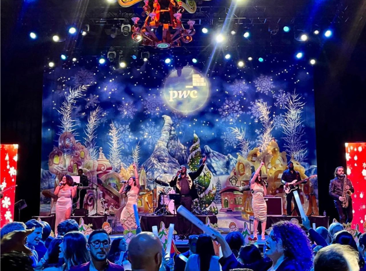 A holiday-themed stage with a snowflake and winter mountain backdrop, Christmas decorations, and a band performing. Audience members are in the foreground.