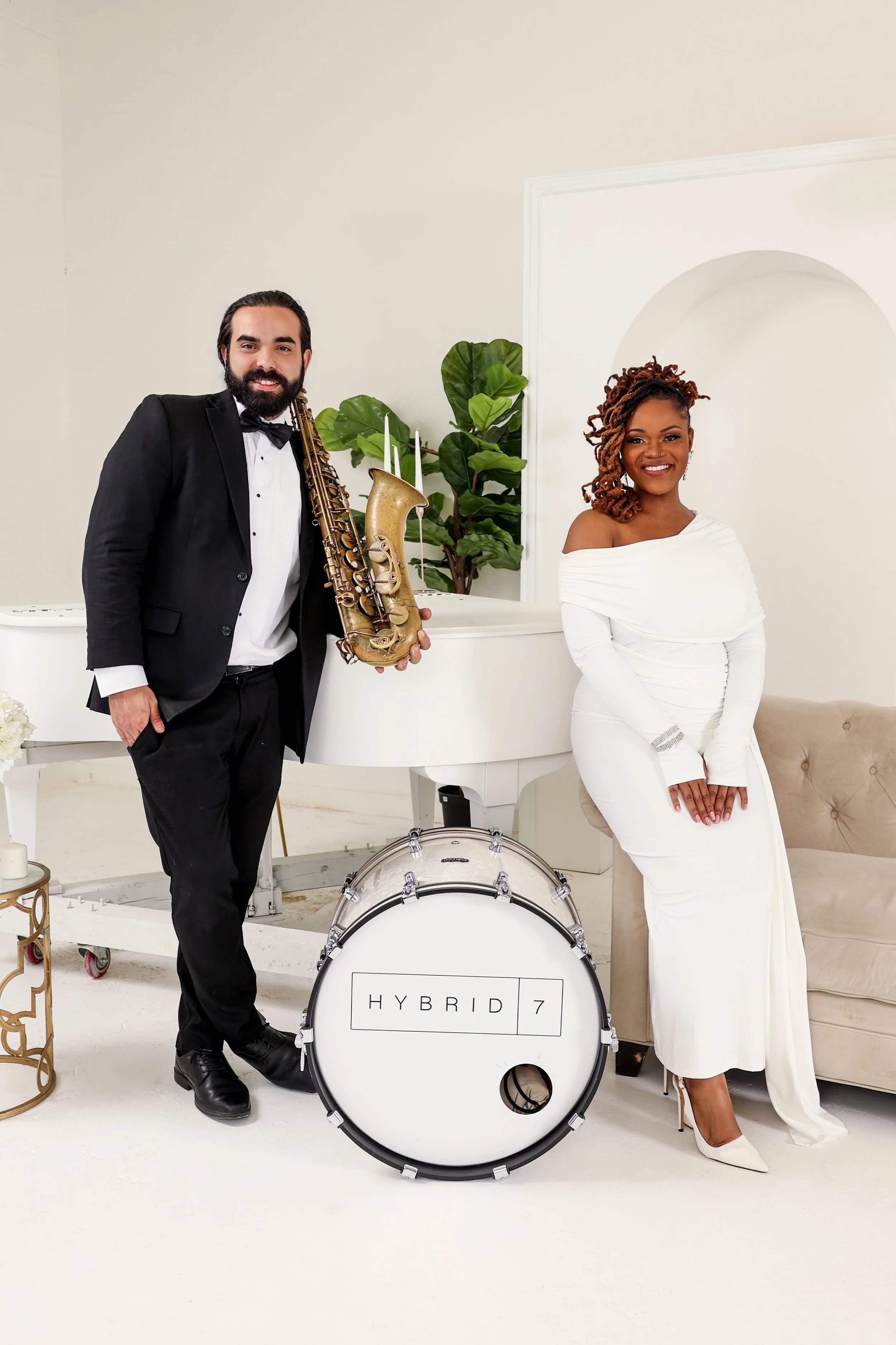 A man in a tuxedo holding a saxophone and a woman in a white dress standing beside a drum with the label "HYBRID 7" in a bright, elegant room with a white grand piano, green plants, and a beige sofa.