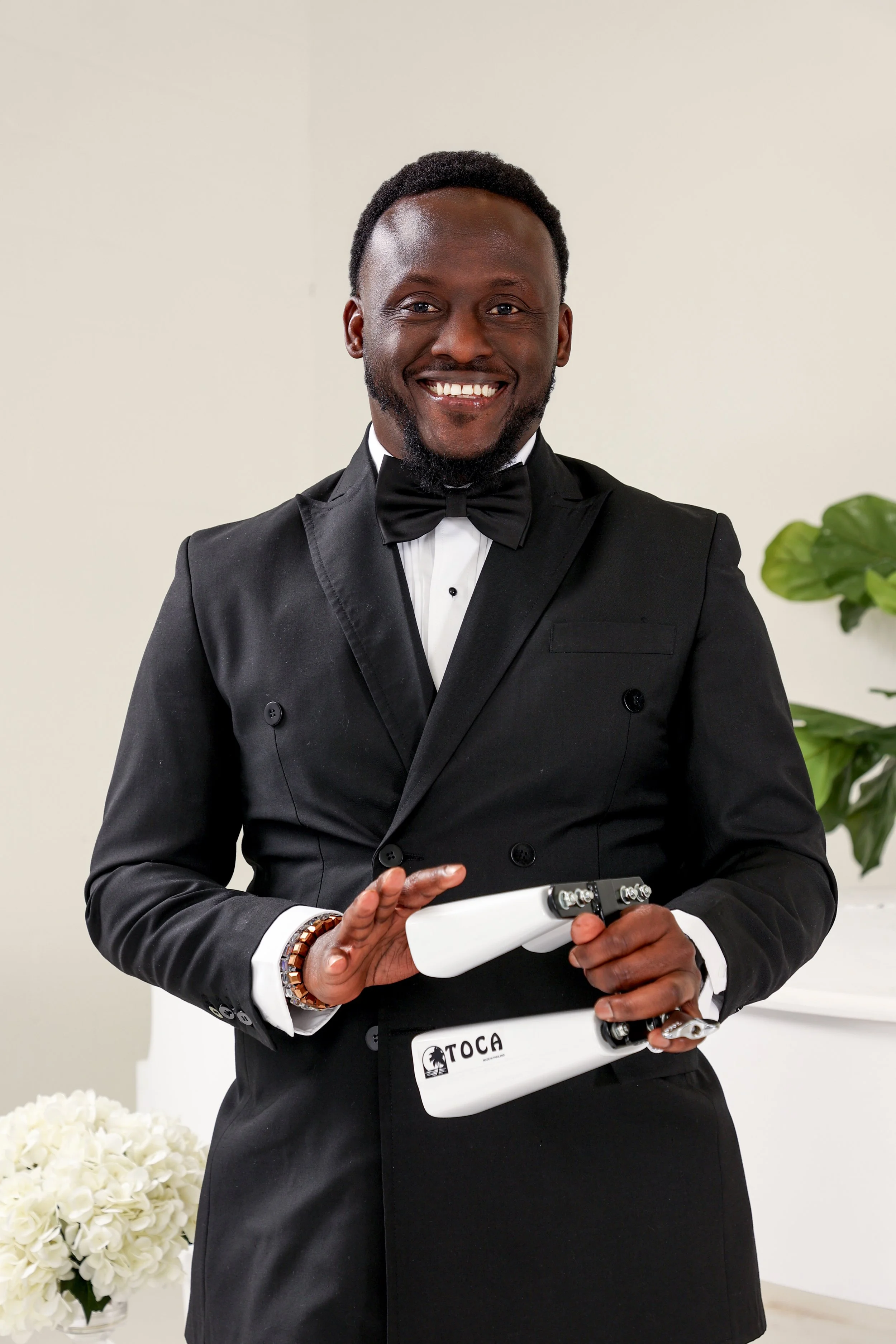 A figurine of a bride and groom dancing on a stack of white plates at a wedding celebration. In the background, there is a pink and white decorated wedding cake and a bouquet of pink and peach roses.