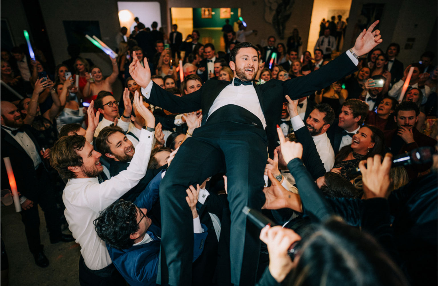 A man in a tuxedo is being lifted by a crowd at a party or celebration, smiling and raising his arms, surrounded by a crowd of well-dressed people.