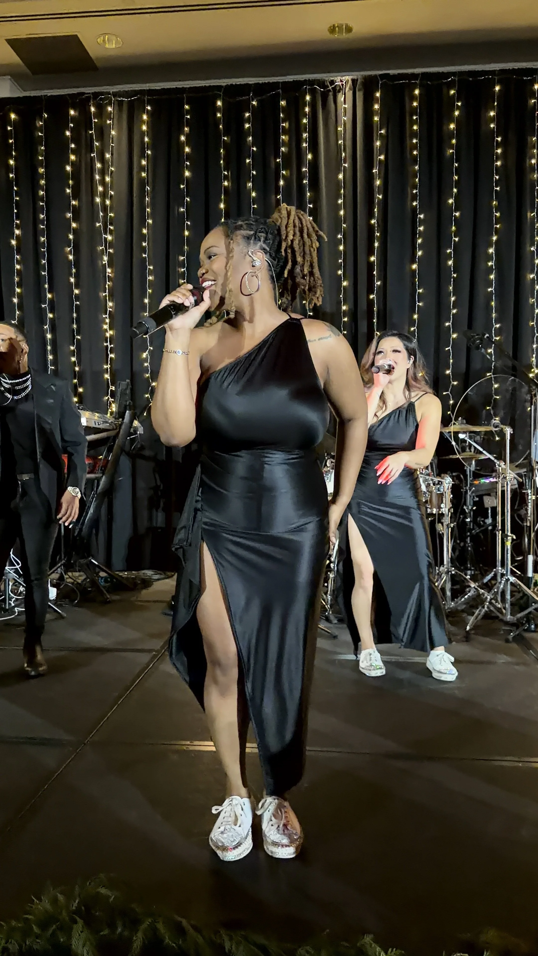 A woman singing into a microphone on stage, wearing a black satin dress with a thigh-high slit and sneakers, with other performers and musical equipment in the background, decorated with string lights.