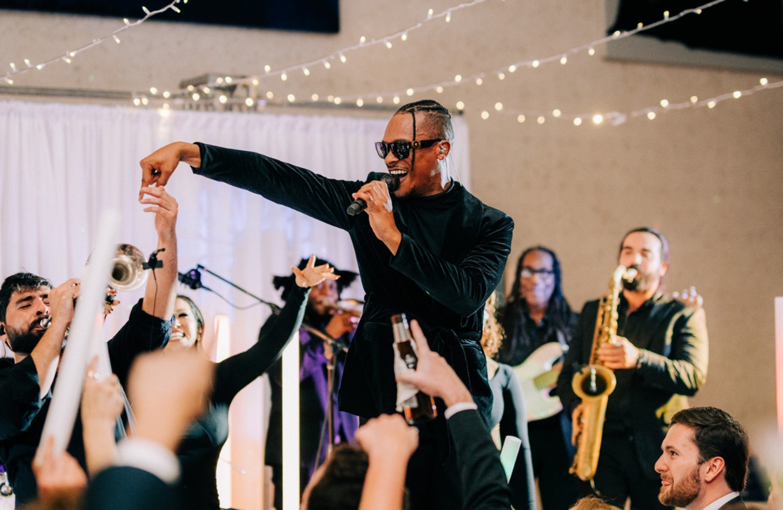 A man with sunglasses singing into a microphone at a lively party or celebration, surrounded by people dancing and playing instruments, with string lights overhead.
