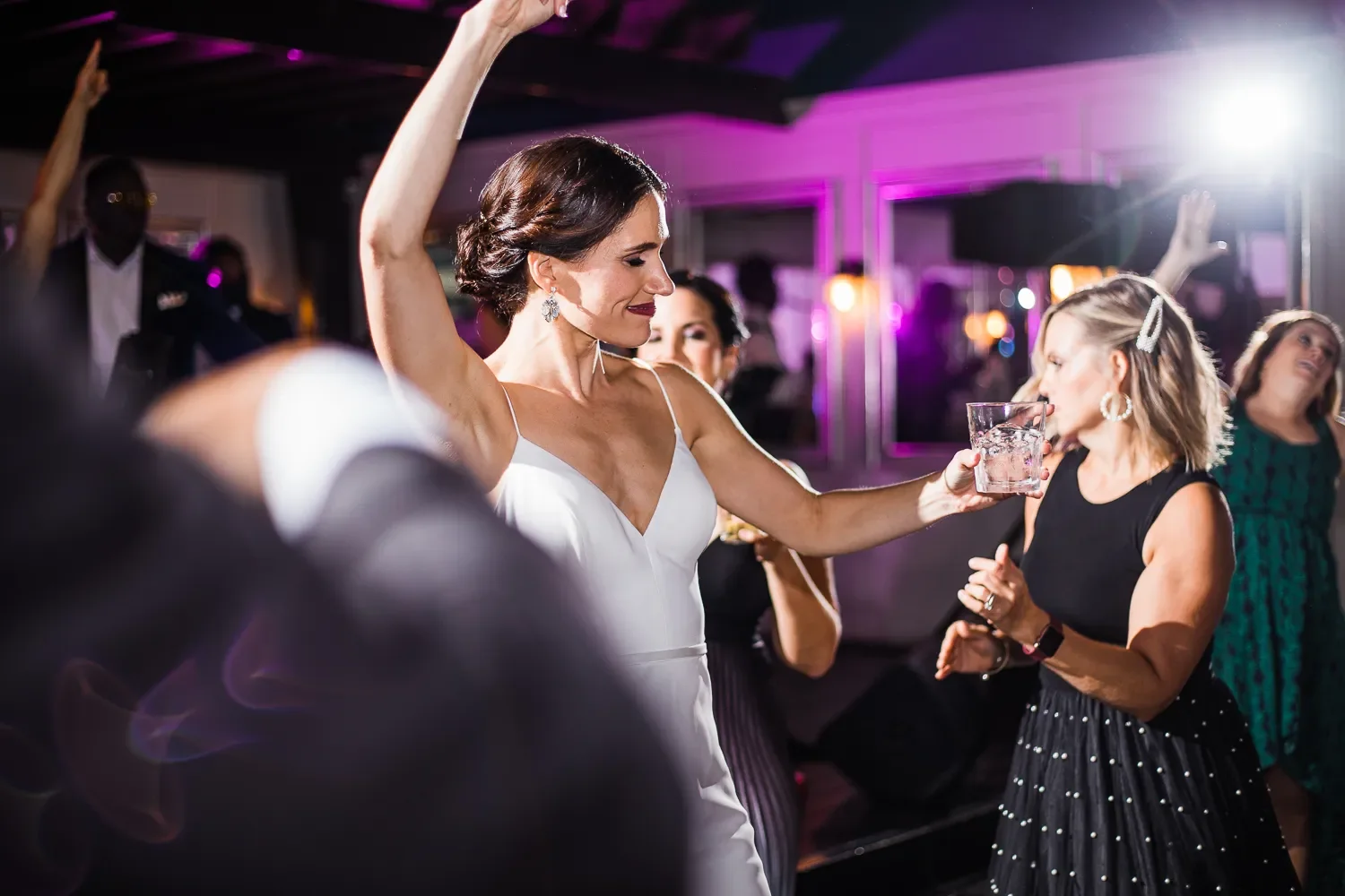 Women dancing and enjoying at a party with purple and pink lighting, some holding drinks.