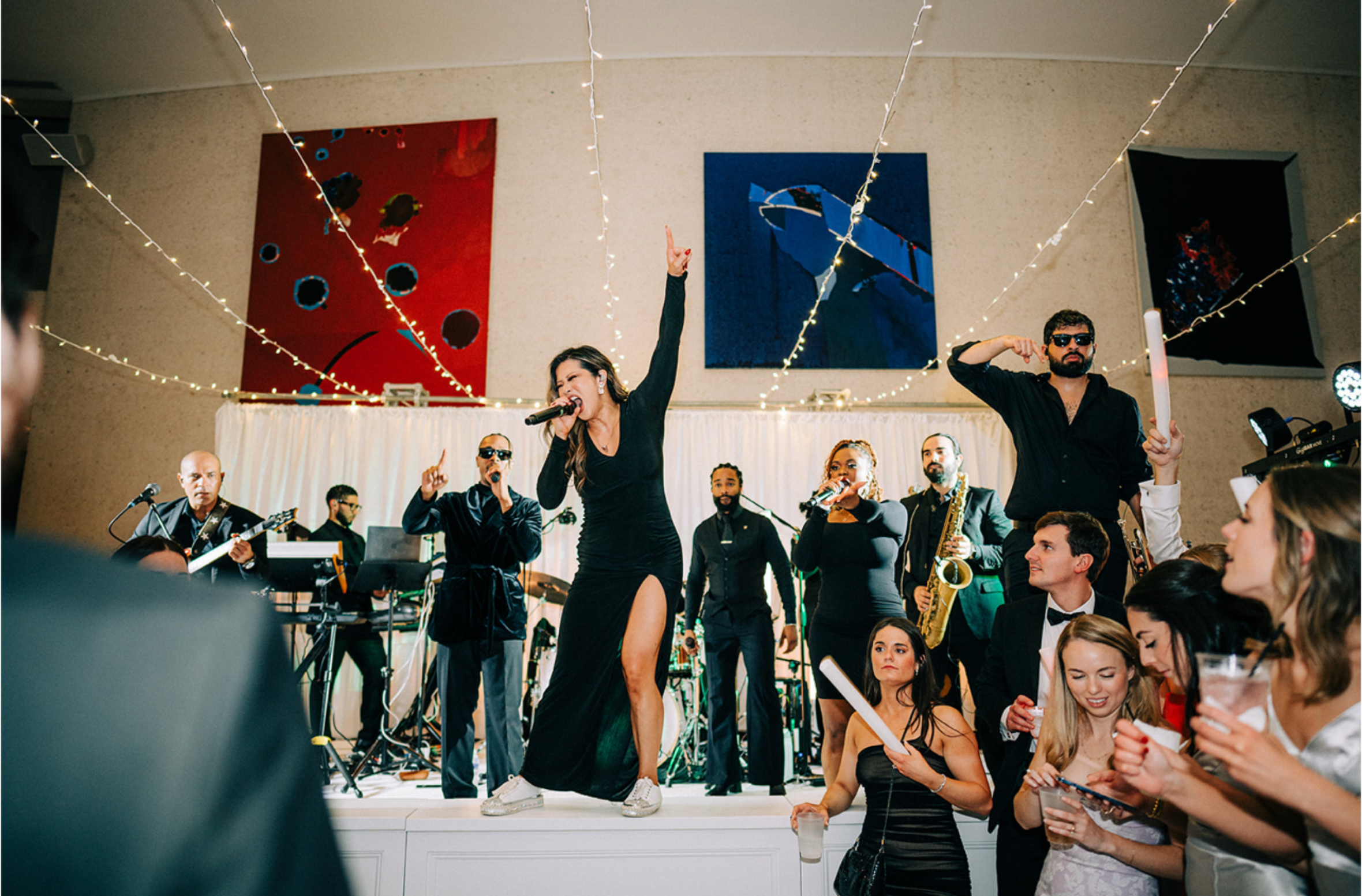 A band performs on stage at a lively party with a woman singing into a microphone, another holding a drink, and people enjoying and dancing. String lights decorate the ceiling, and guests are dressed in formal and stylish attire.