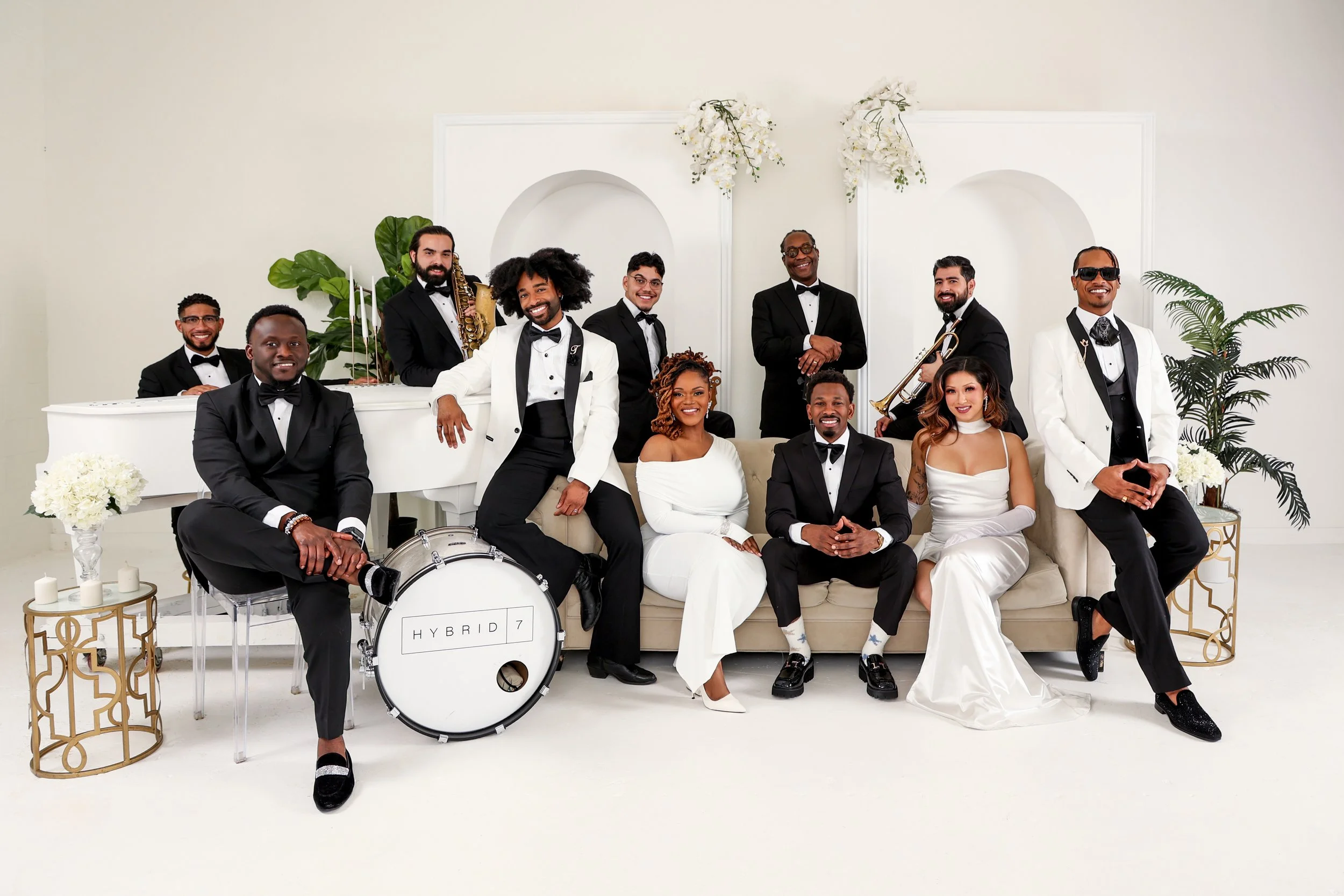 Group of eleven people in formal attire posing in a bright, elegant room with white walls and white decor, including a white grand piano and a sofa, with some holding musical instruments.