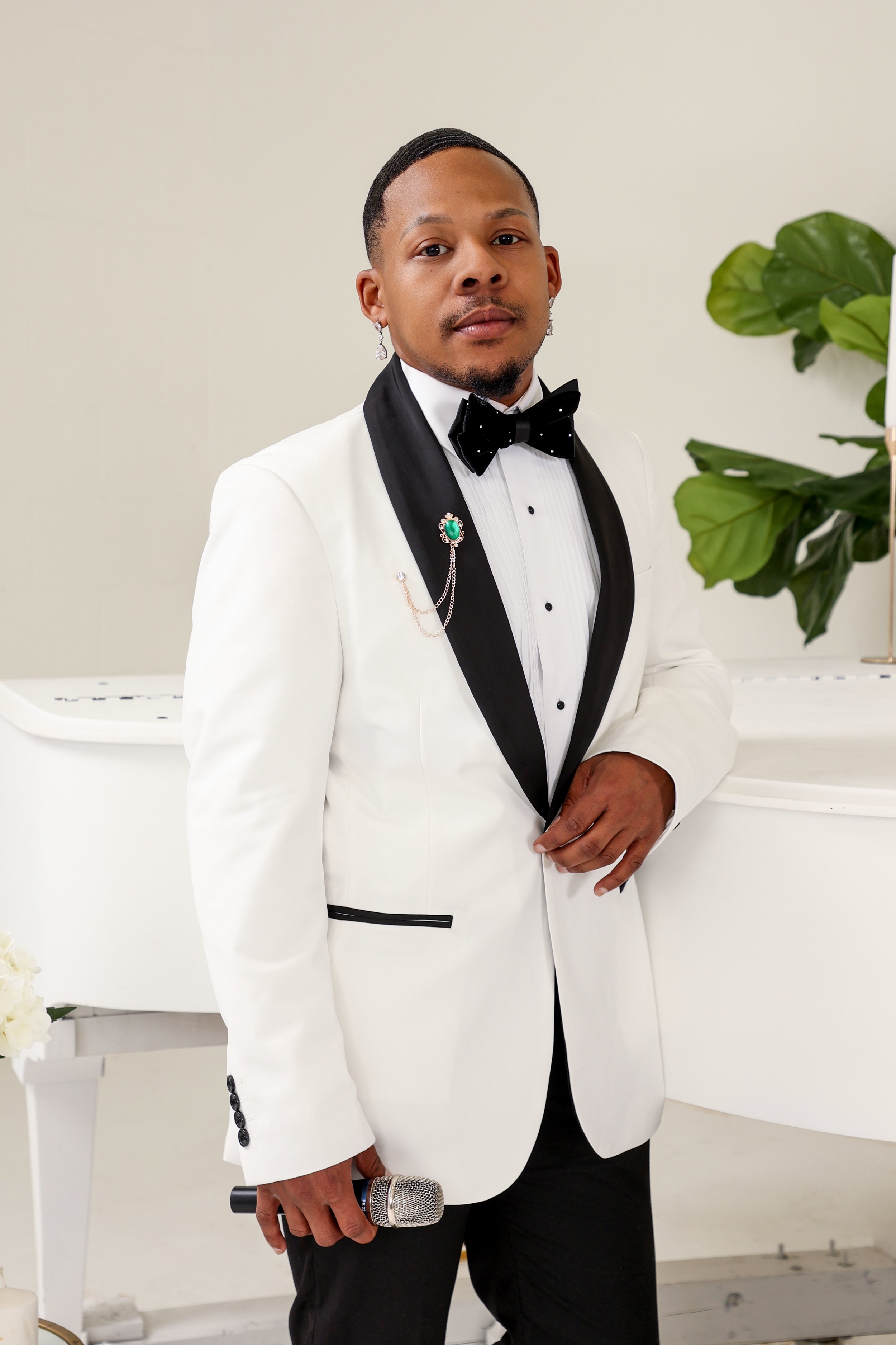 A man dressed in a white tuxedo with black lapels, a black bow tie, and black pants. He is holding a microphone and standing indoors near a white grand piano and a large green plant.