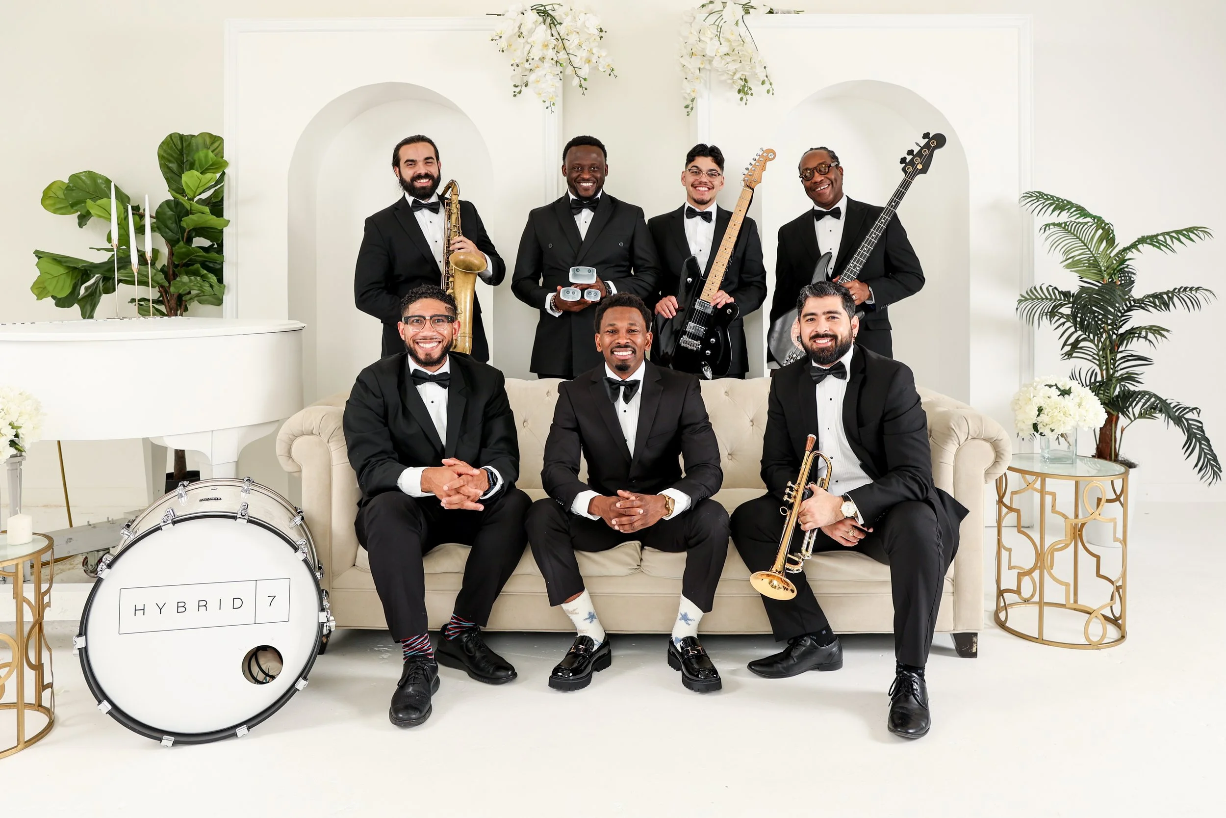 A band of seven men dressed in black tuxedos with bow ties, posing in a bright, elegant room with white walls and decorative plants. They are holding various musical instruments, including saxophone, electric guitar, bass guitar, trumpet, and drums, 